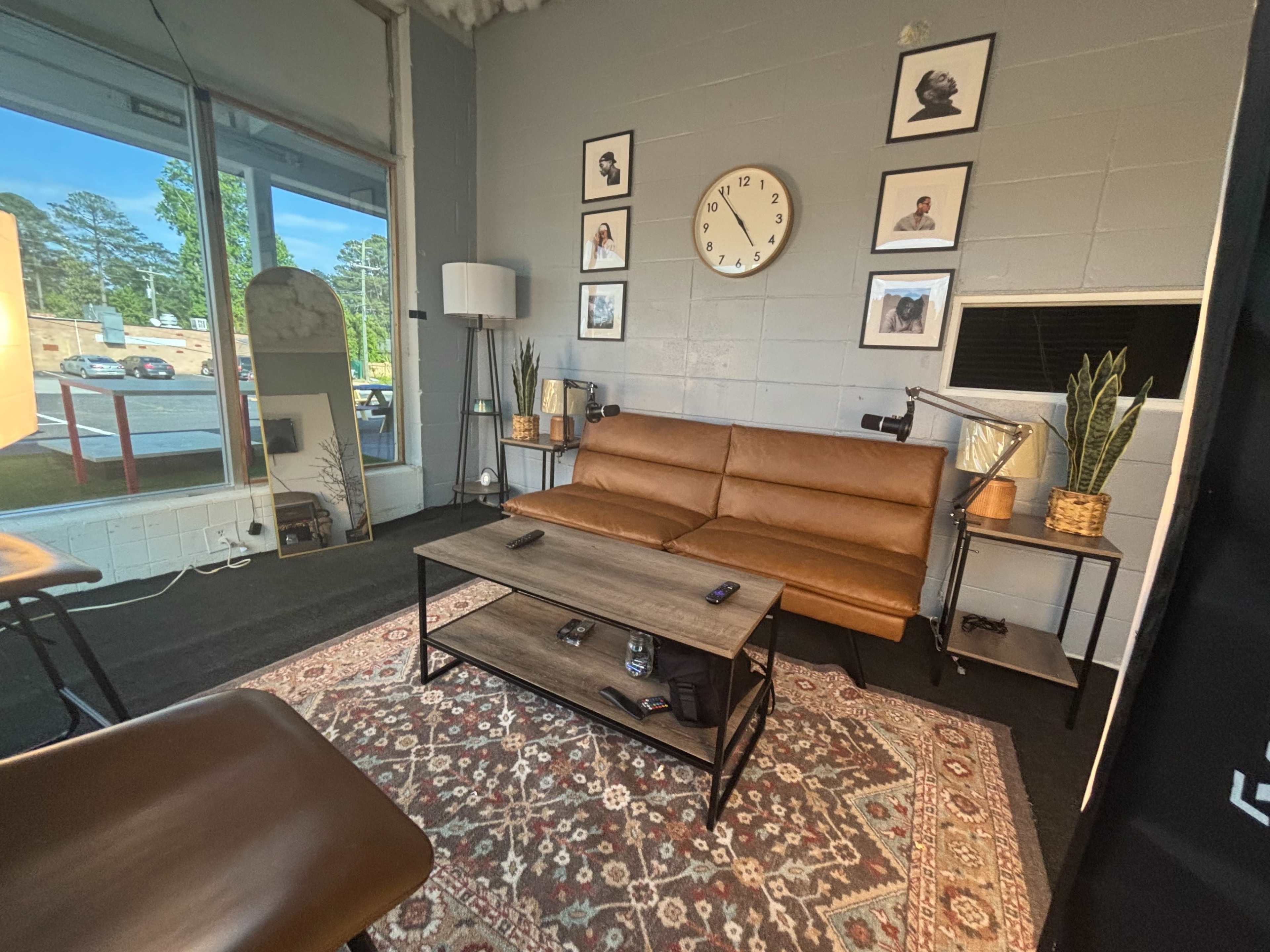 The image shows a modern interior space with a brown leather couch, a wooden coffee table, decorative plants, a round wall clock, and framed pictures on the wall.