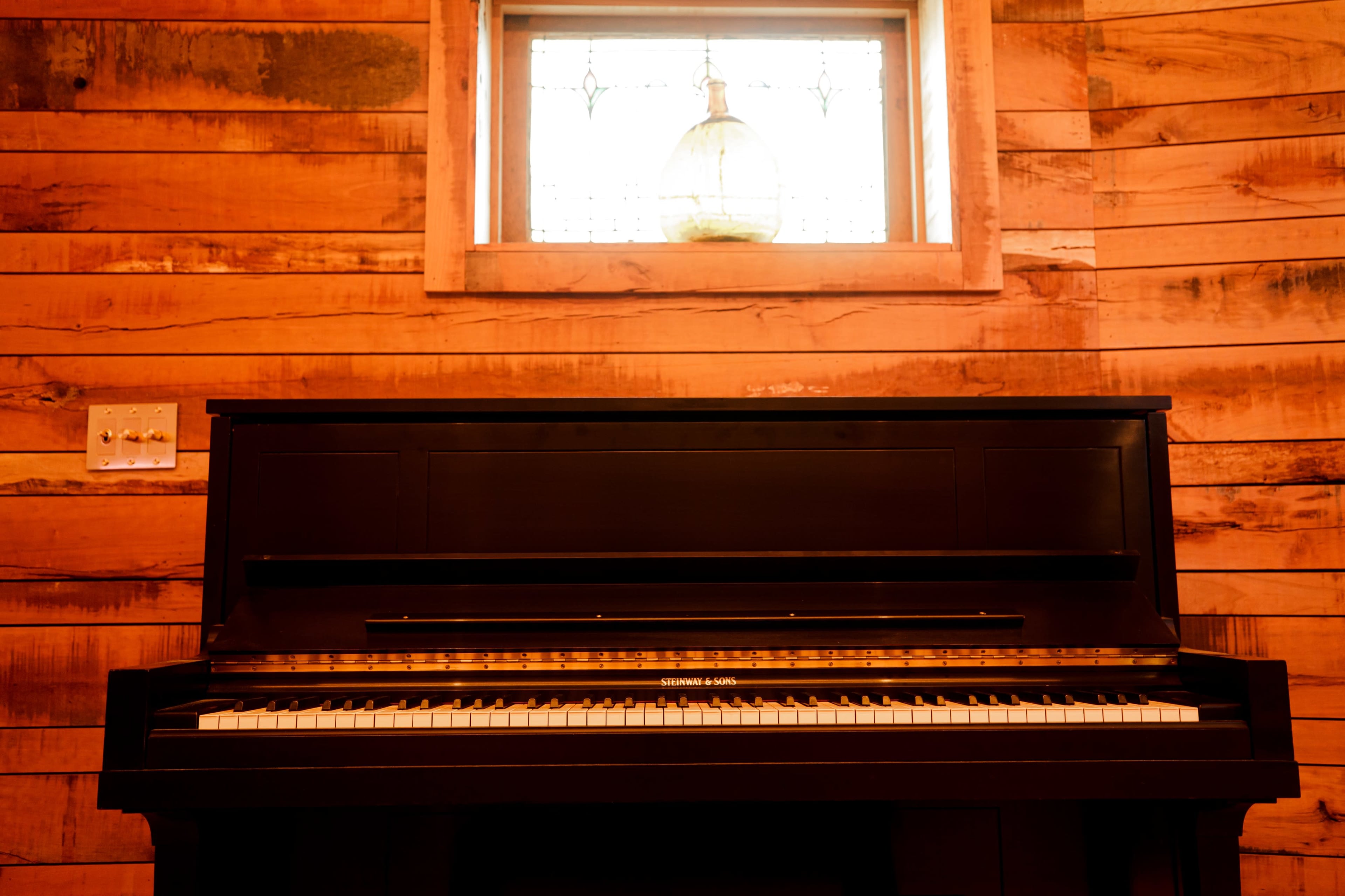 A black piano is positioned in front of a window with a round glass pendant light, set against wooden walls.