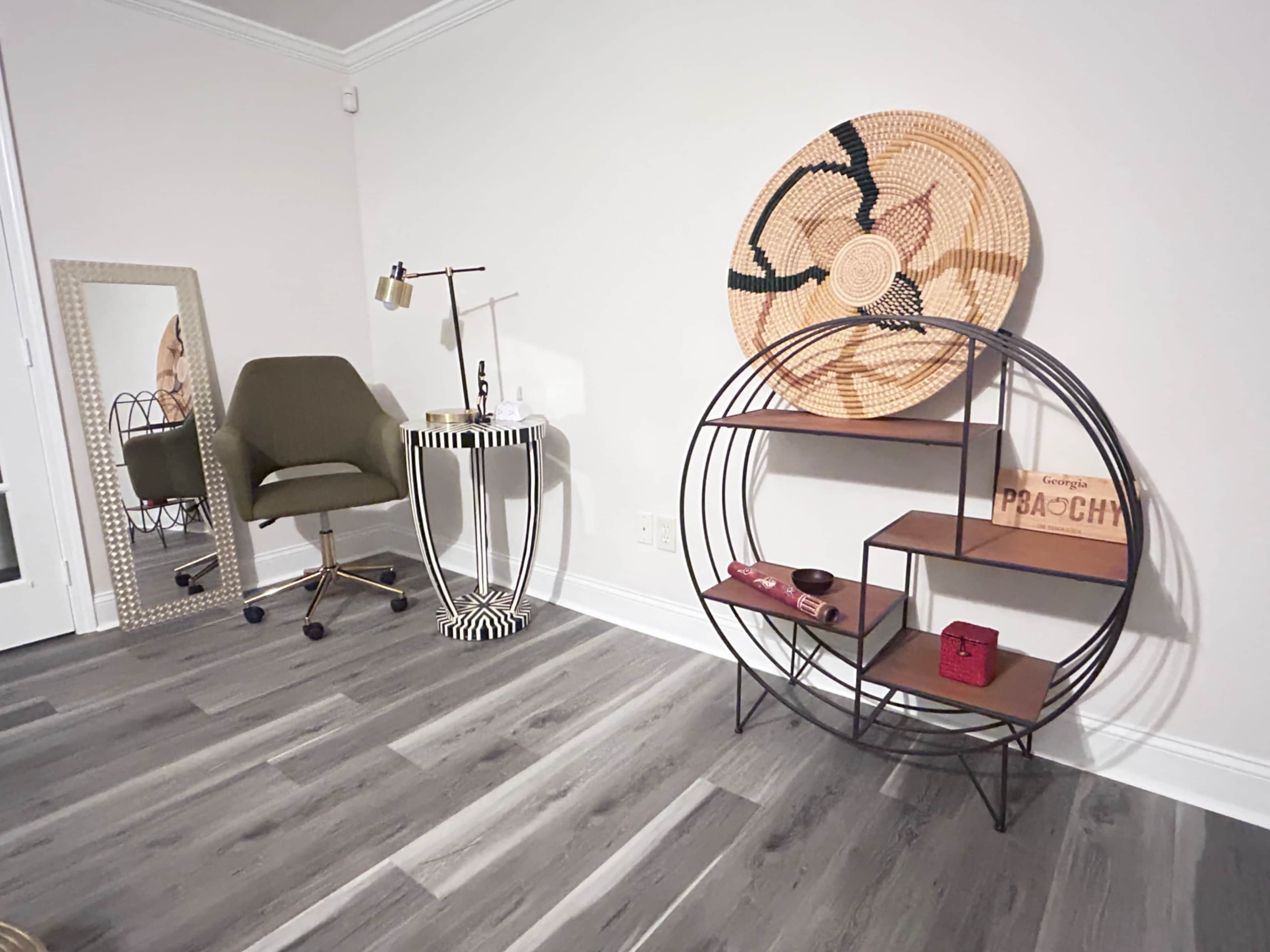 The room features a modern design with a green chair, a round shelf with wooden and metal elements, a patterned table lamp, and decorative wall art.
