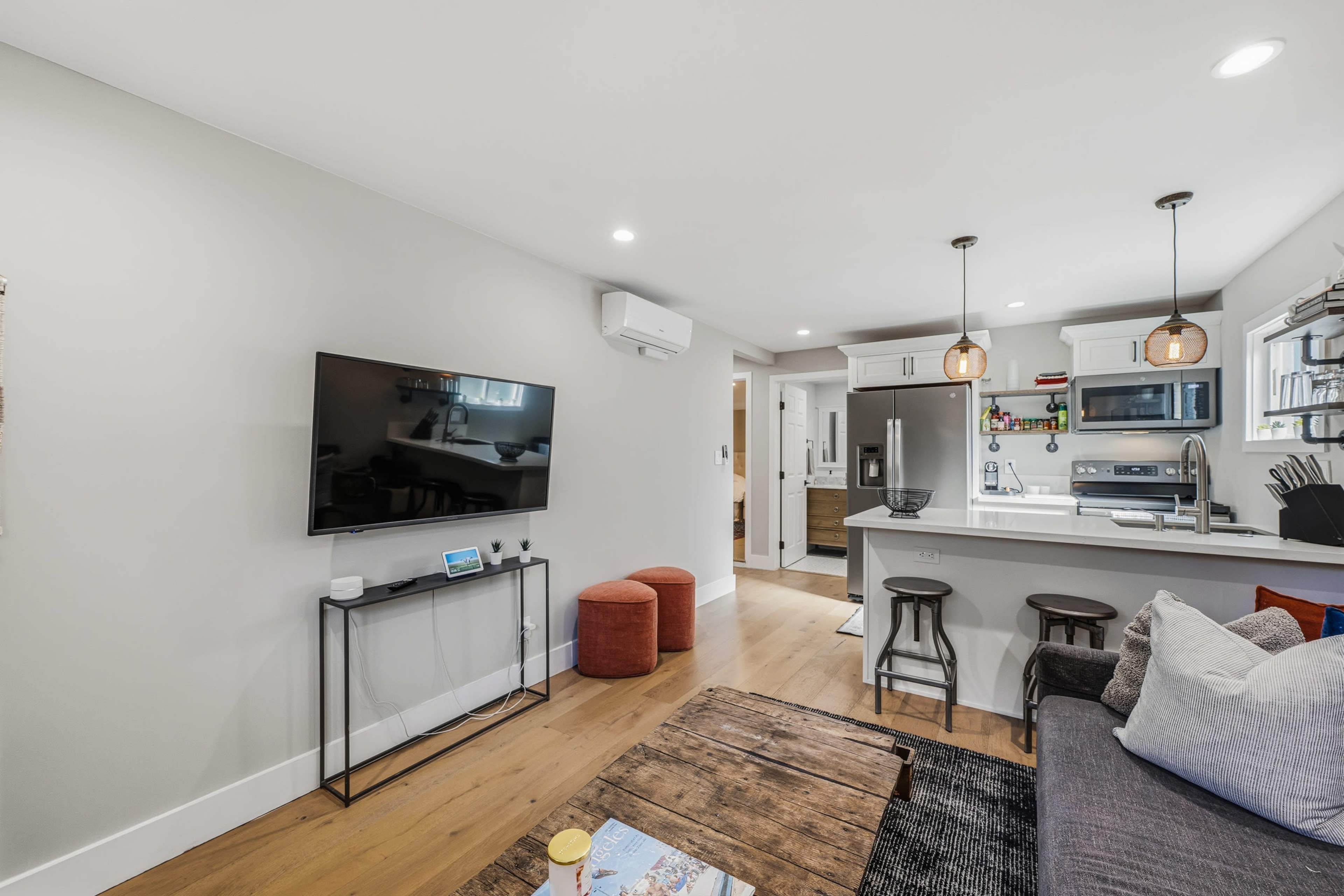 The image shows a modern living room with a television mounted on the wall, a kitchen area in the background, and a sectional sofa, accompanied by a wooden coffee table and a couple of ottomans.