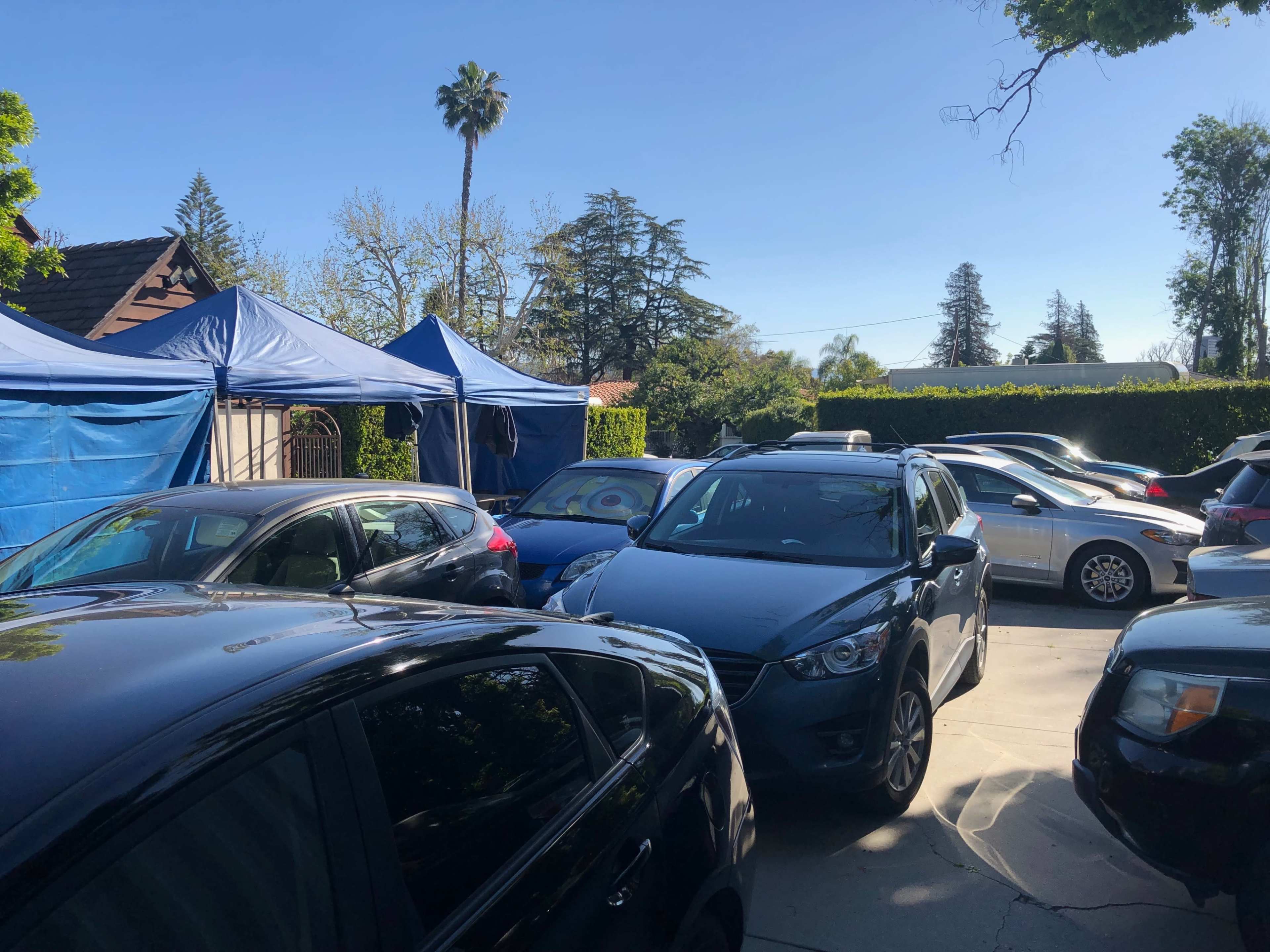 The image shows a crowded parking area filled with various parked cars and tents under a clear blue sky.