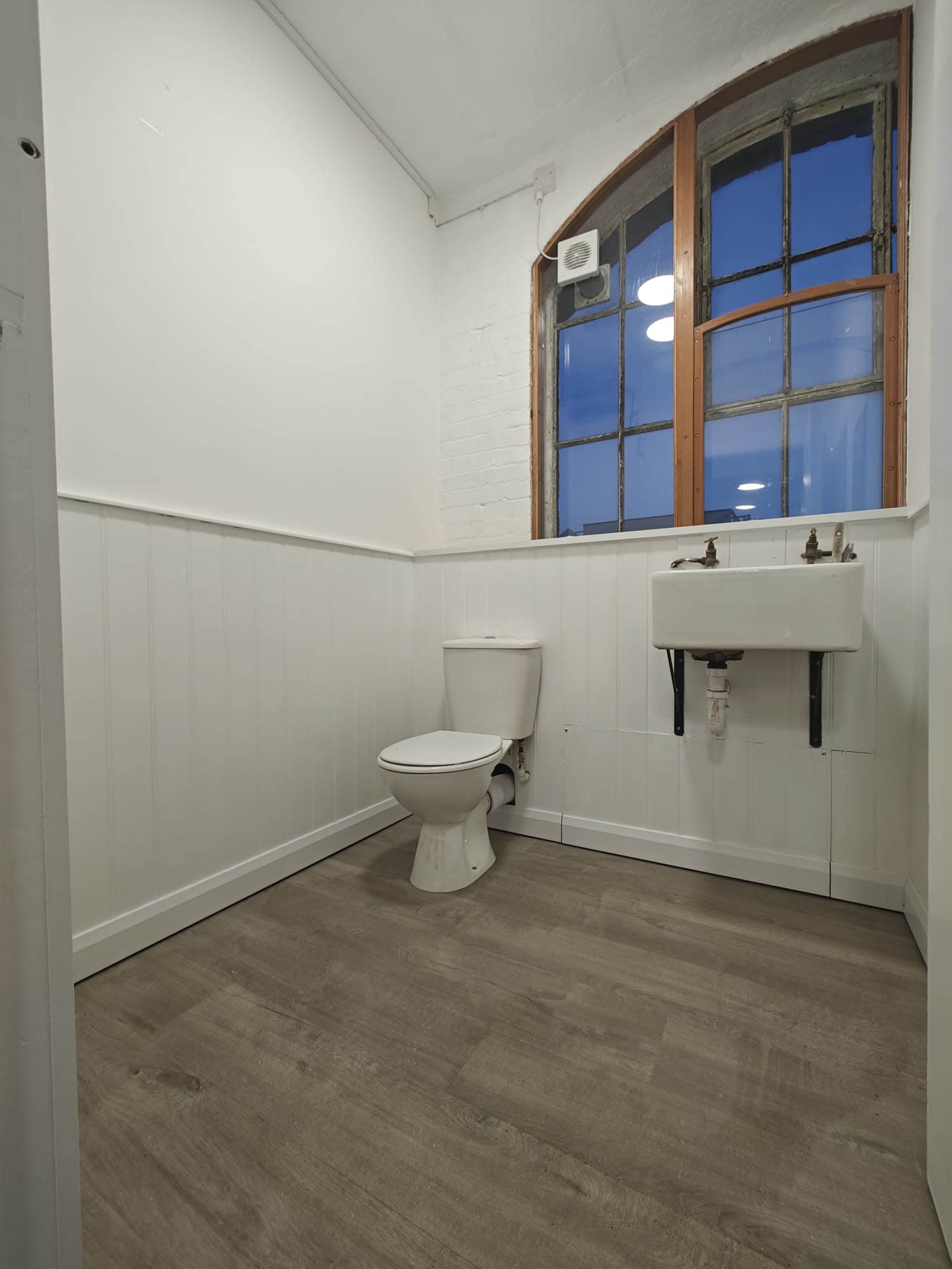 The image shows a clean, well-lit bathroom featuring a toilet, a small sink, and a window with a view of the sky.