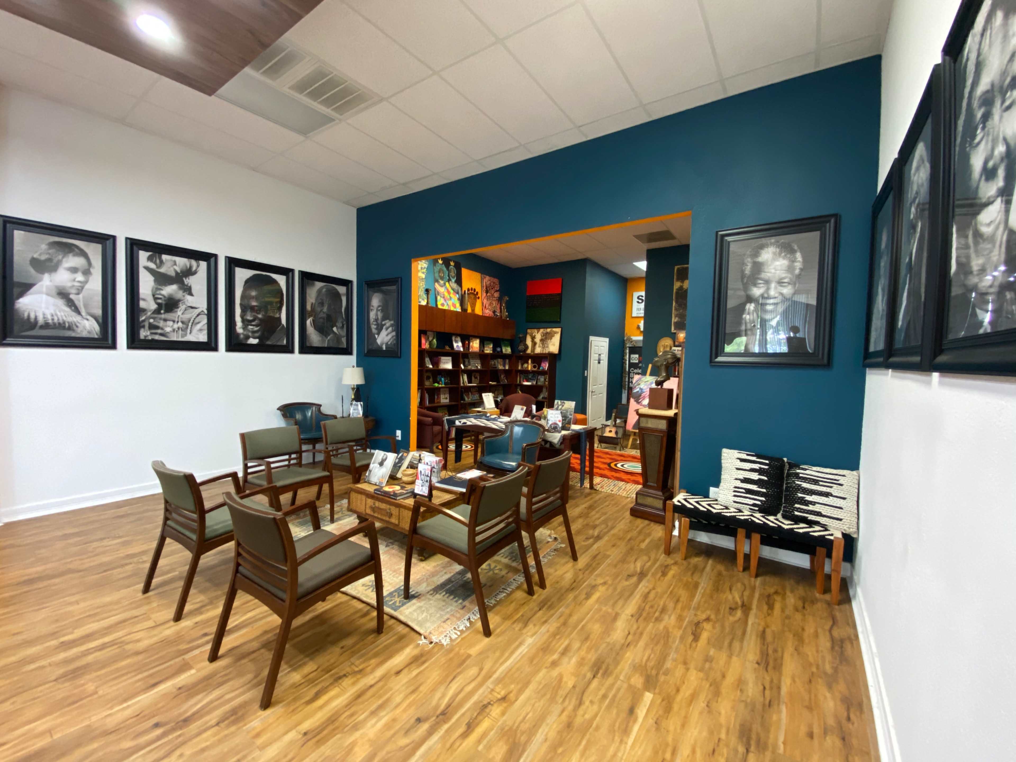 A cozy room with wooden flooring, featuring a seating area with chairs and tables, surrounded by large black and white portraits on the walls, and a bookcase filled with items in the background.