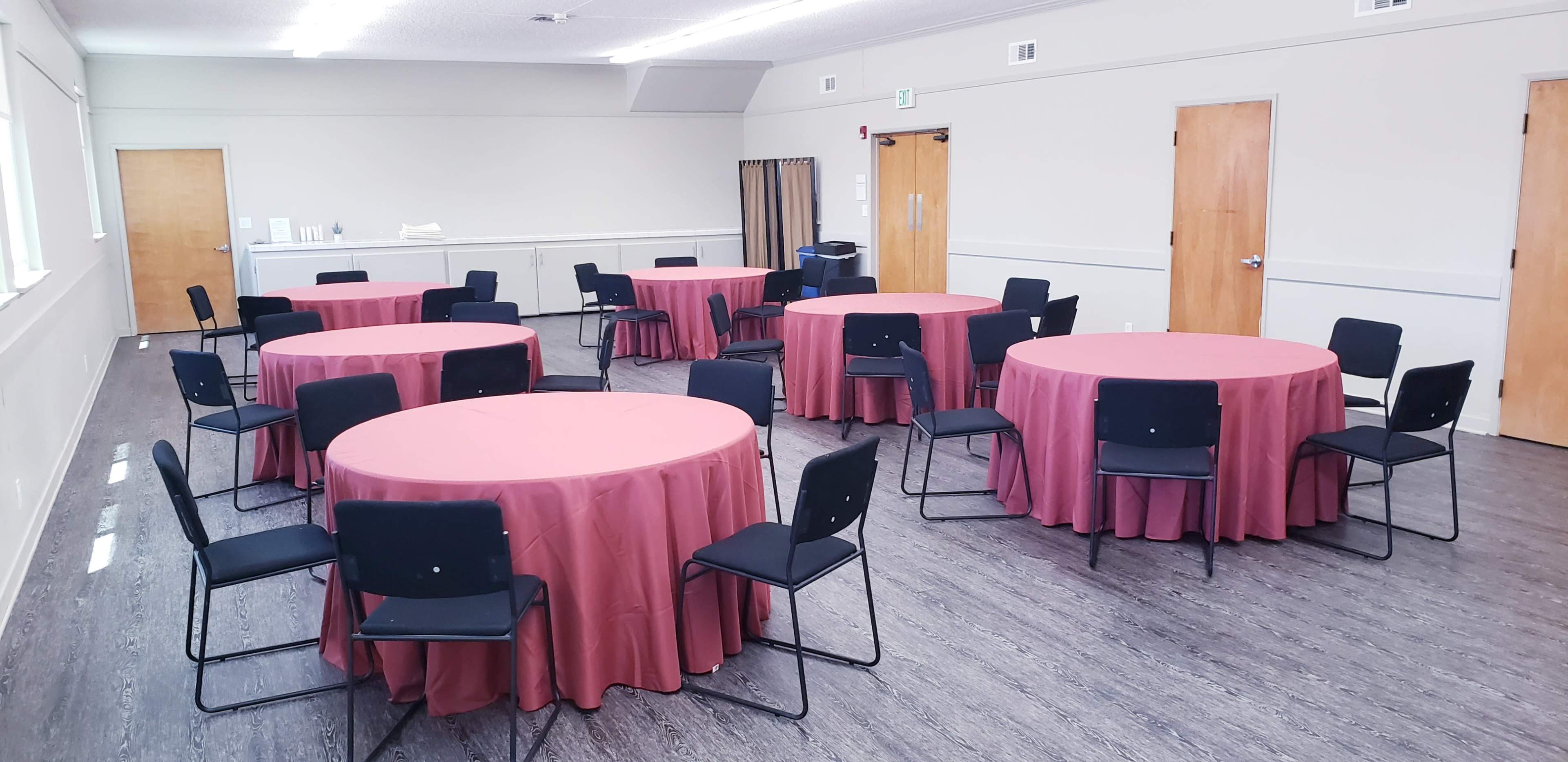 A large room contains six round tables covered with maroon tablecloths, each surrounded by black chairs.