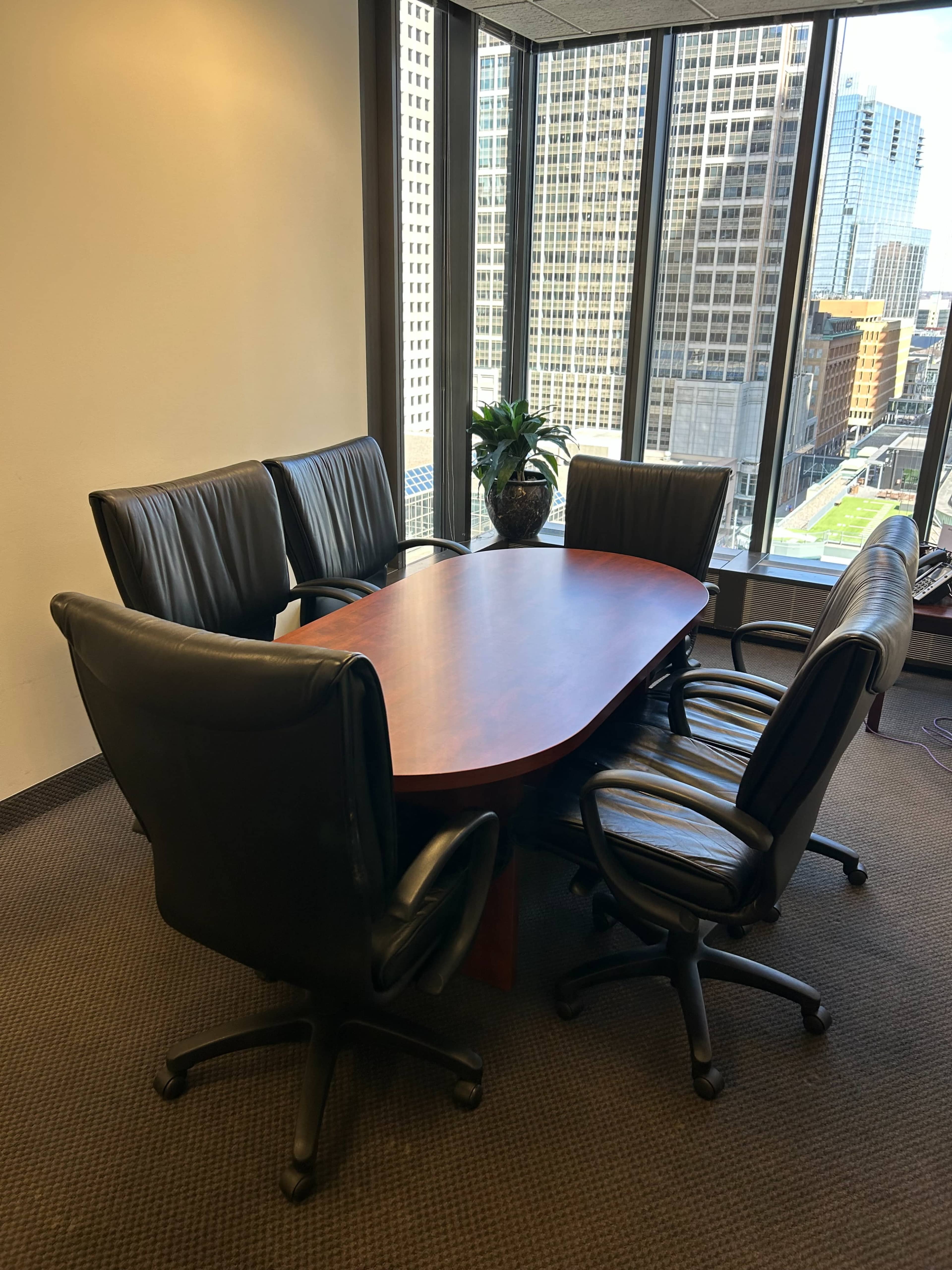 A conference room features a large oval table surrounded by six black leather chairs, with large windows displaying a cityscape in the background.