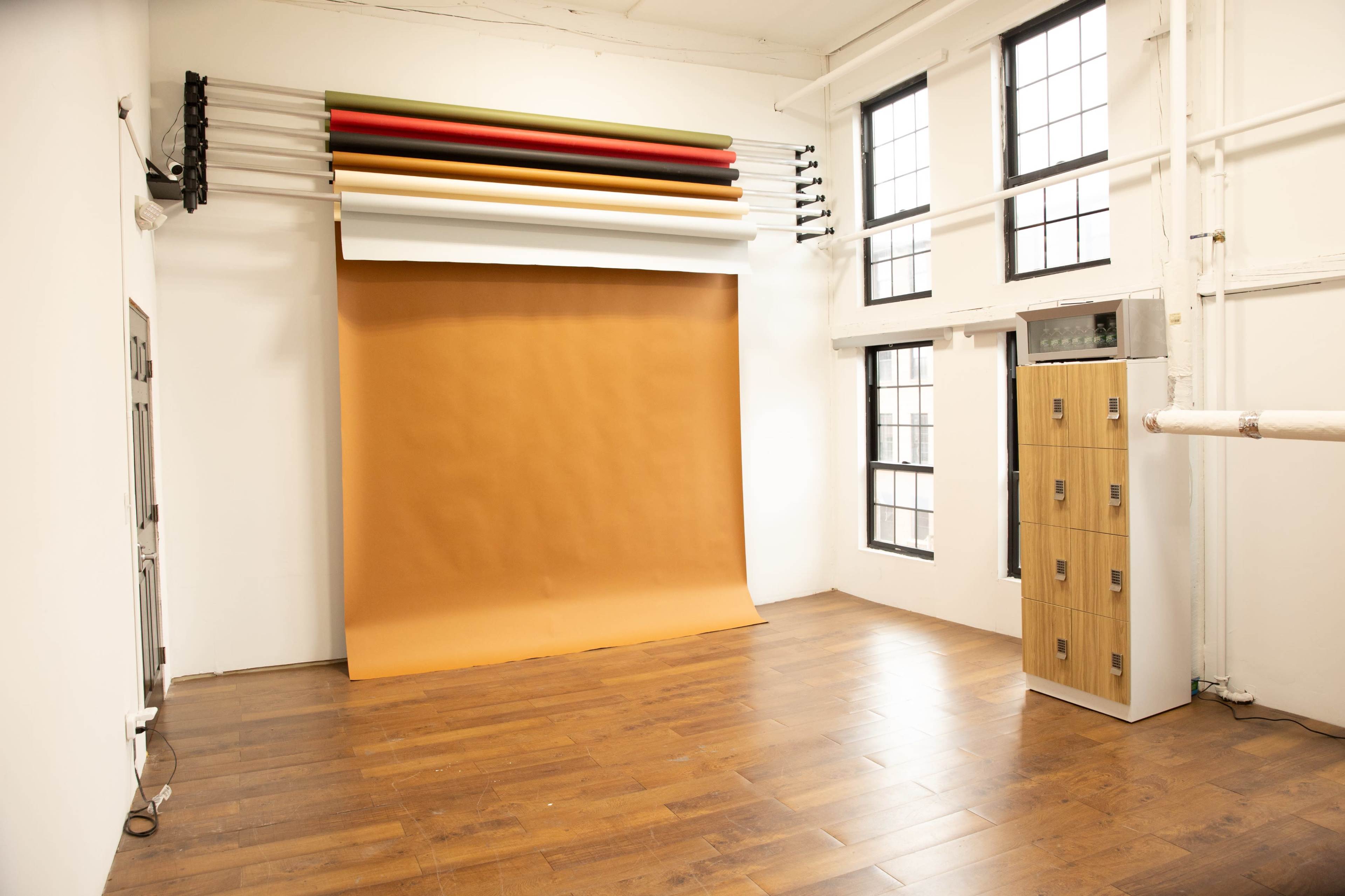 The image shows a spacious room with a brown backdrop roll, large windows, and a wooden floor, alongside a cabinet with multiple drawers.