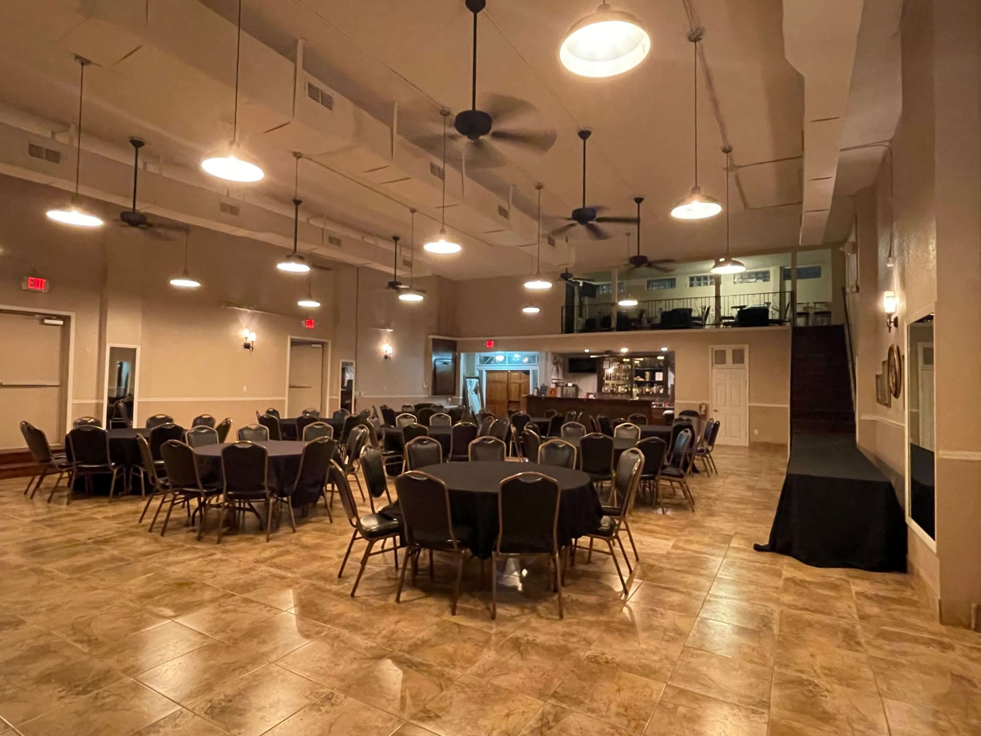 The image shows a banquet hall with several round tables covered in black tablecloths, set within a spacious room featuring ceiling fans and a bar area in the background.