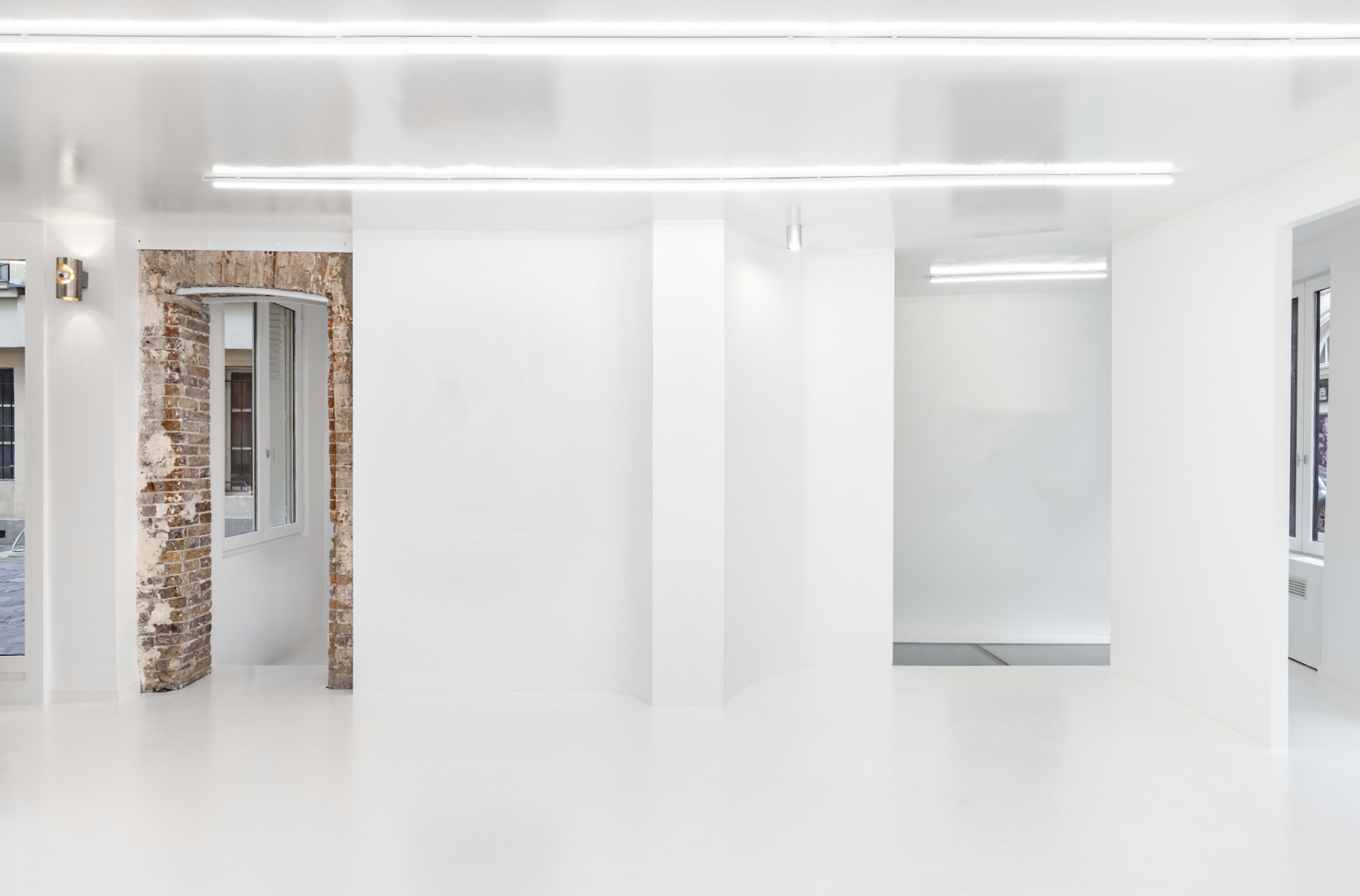 The image shows a minimalist interior space with white walls and a polished floor, featuring a section of exposed brick and large windows.