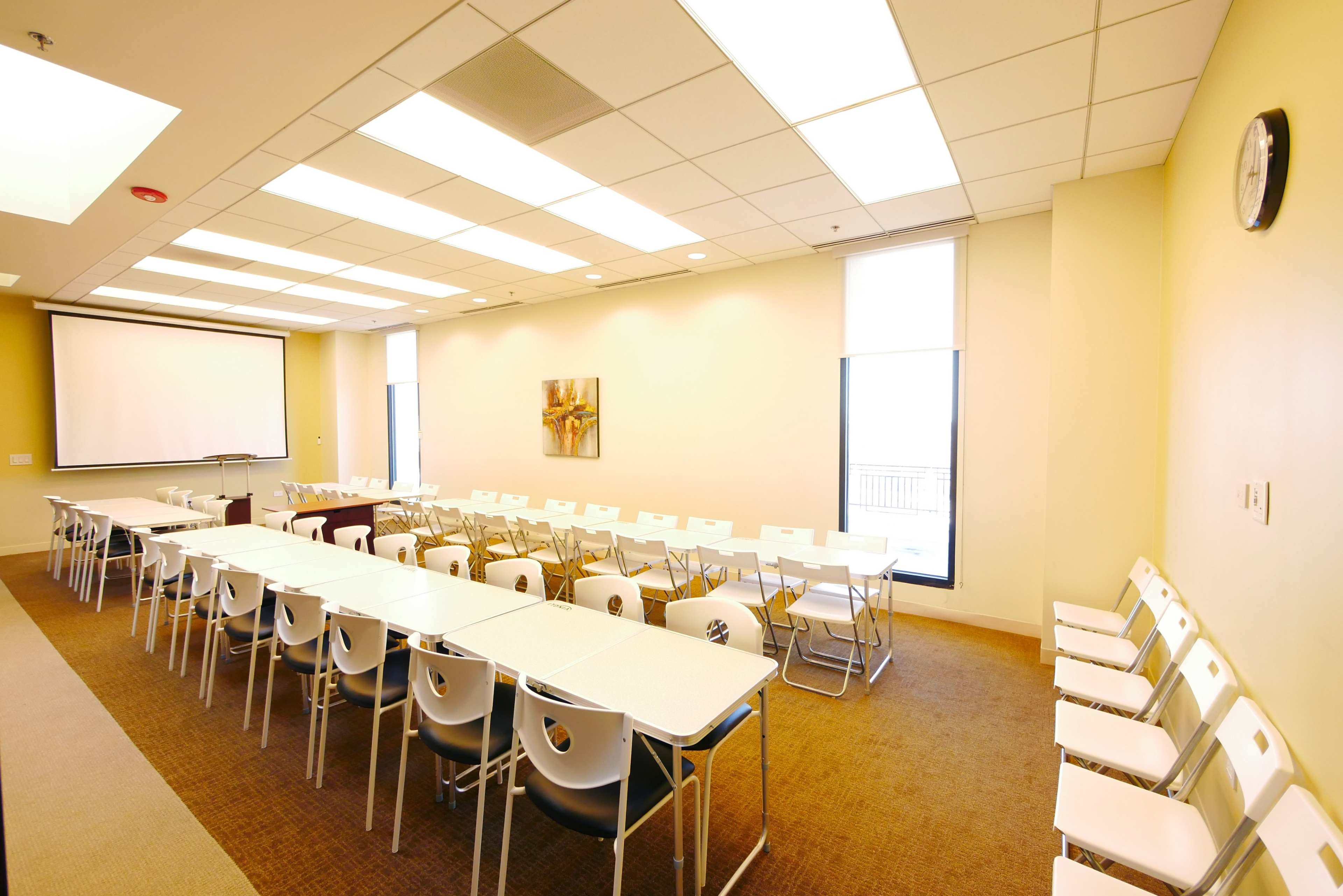 The image shows a well-lit meeting room with multiple tables arranged in a U-shape and rows of white chairs along the walls.