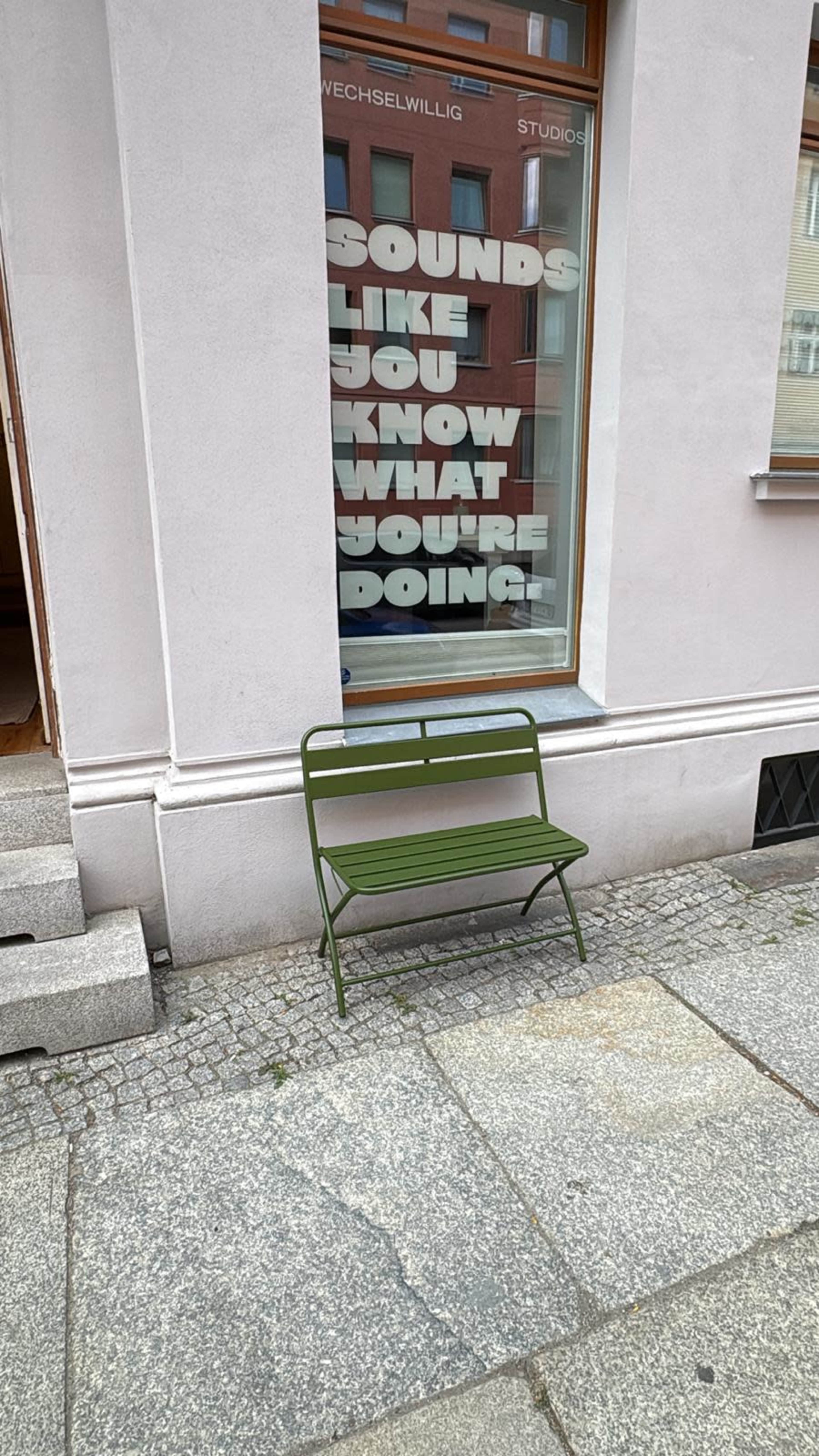 A green metal bench is positioned next to a building with a large window displaying the text "SOUNDS LIKE YOU KNOW WHAT YOU'RE DOING!"