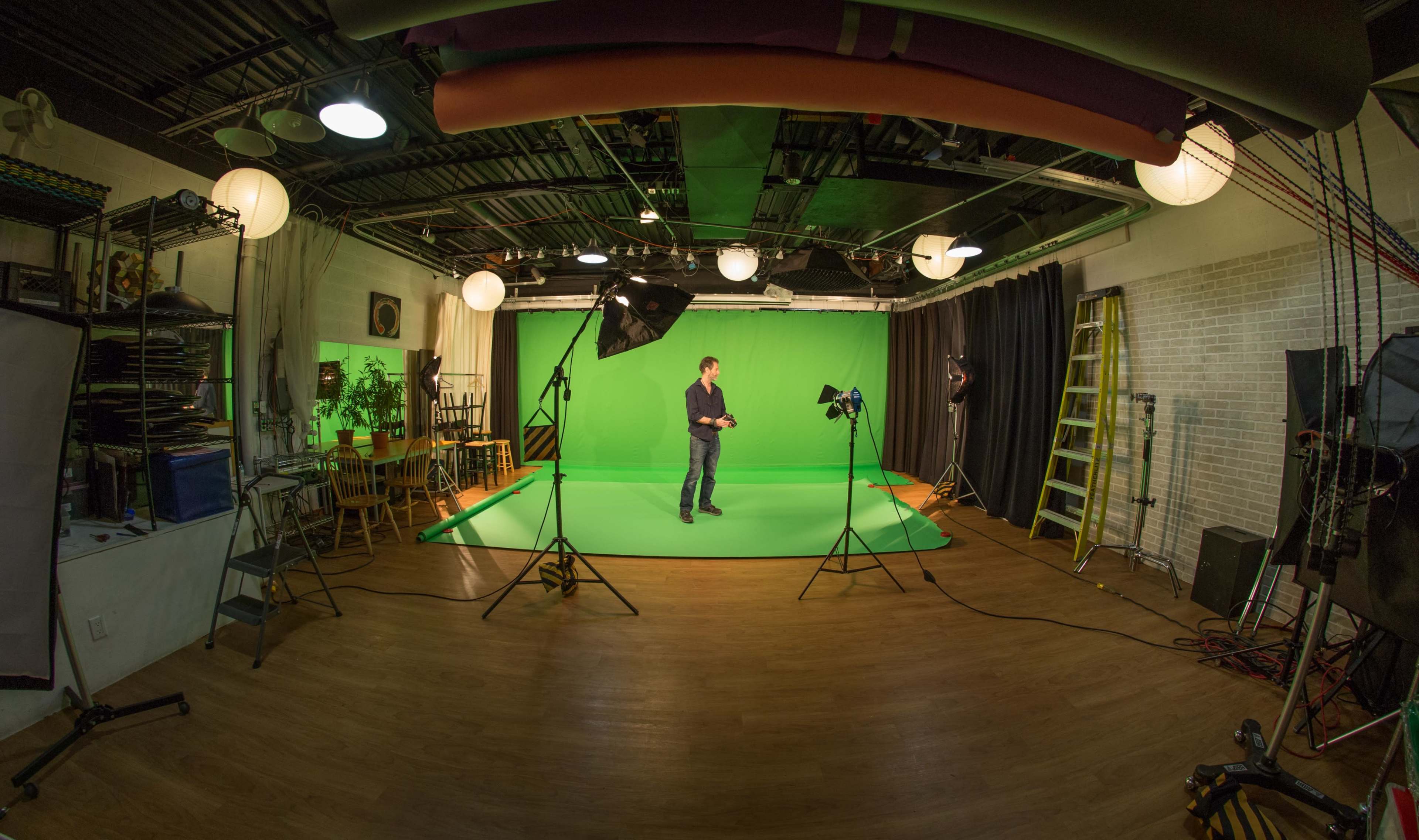 A man stands in a studio with green screens, studio lights, and various equipment arranged around him.