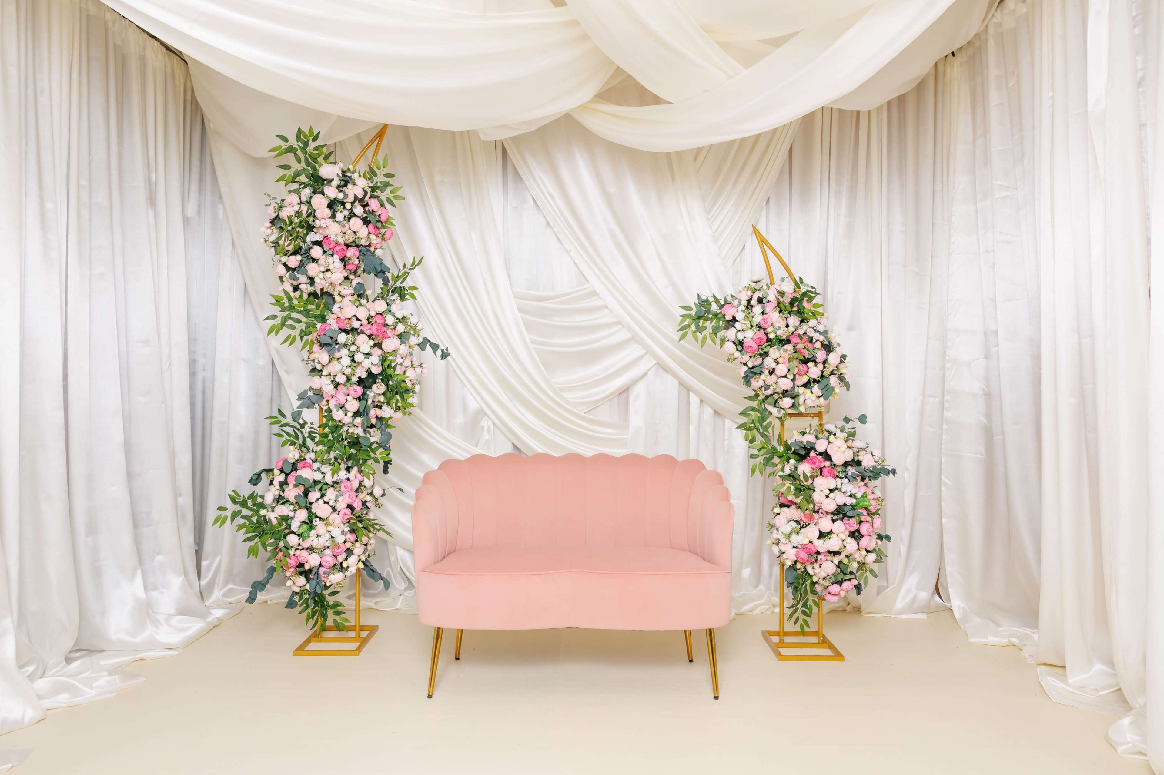 The image features a pink scalloped couch positioned in front of a backdrop of draped white fabric and floral arrangements.