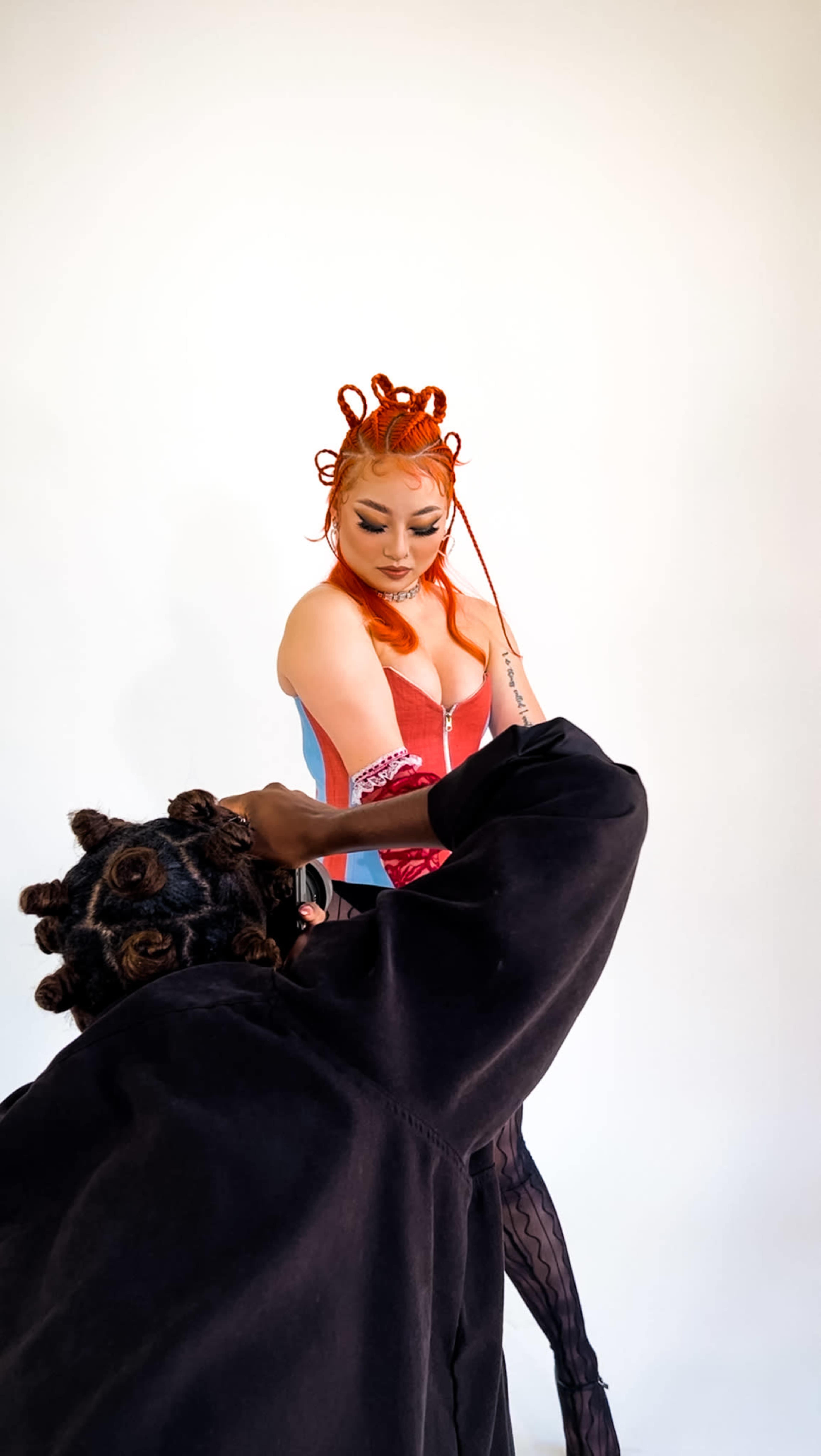 A person with orange hair styled in knots poses while another person adjusts their outfit from behind.