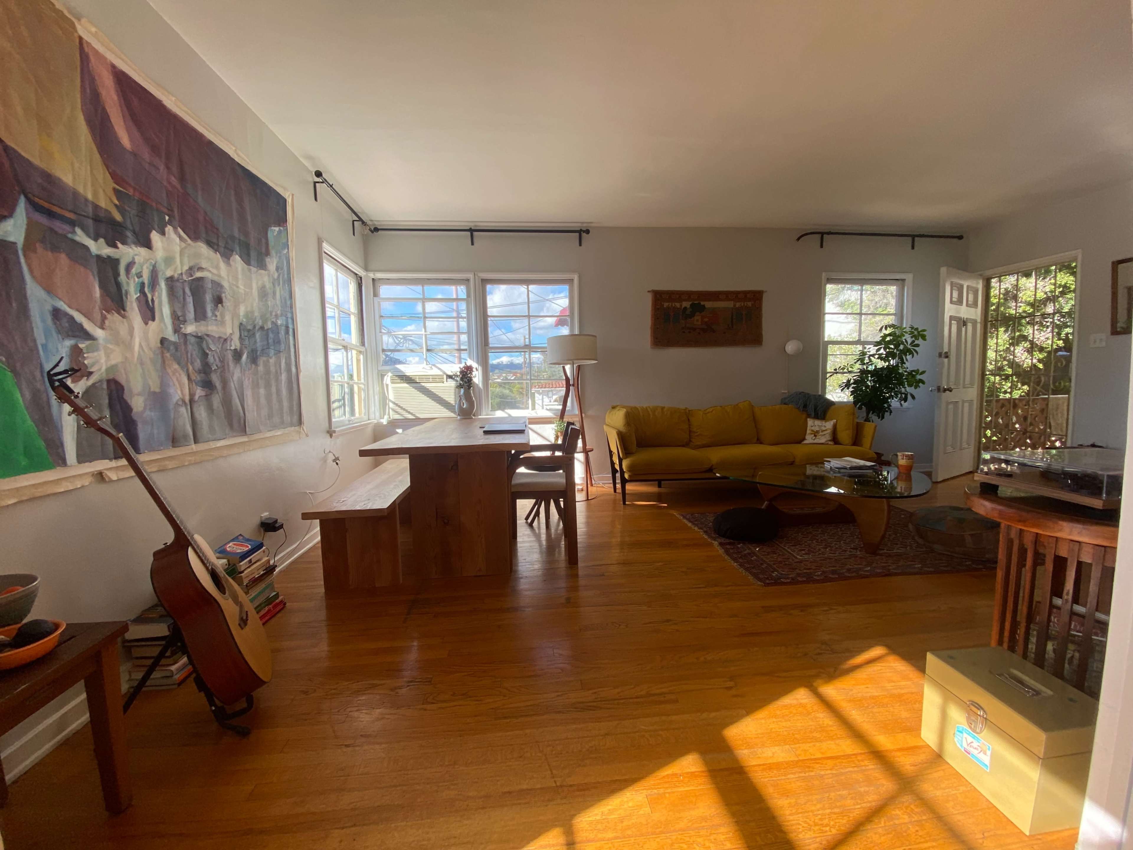 The image shows a bright, open living room space with a yellow sofa, a wooden dining table, and large windows letting in natural light.