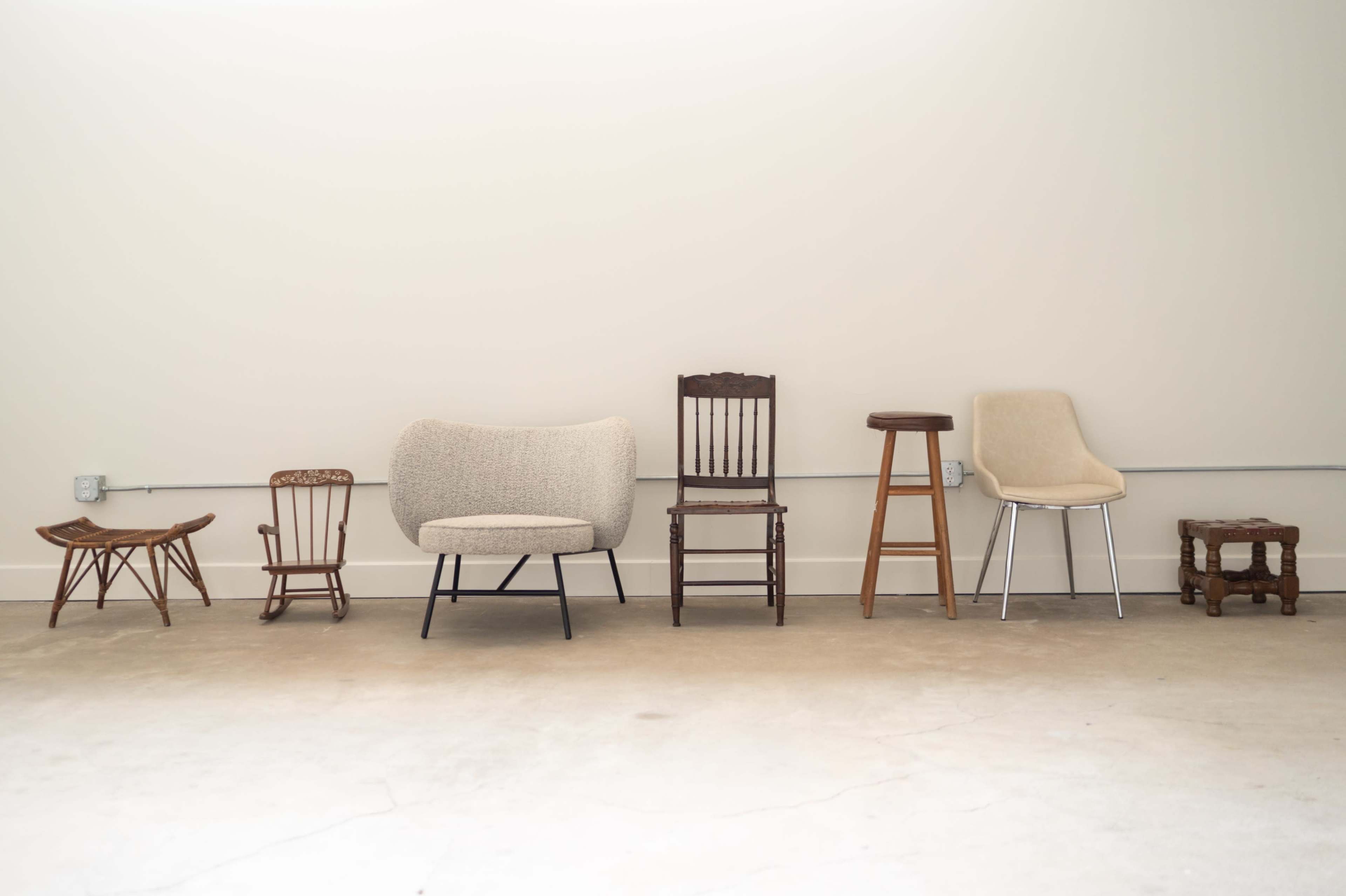The image shows a row of various chairs arranged against a plain wall, with a concrete floor beneath them.