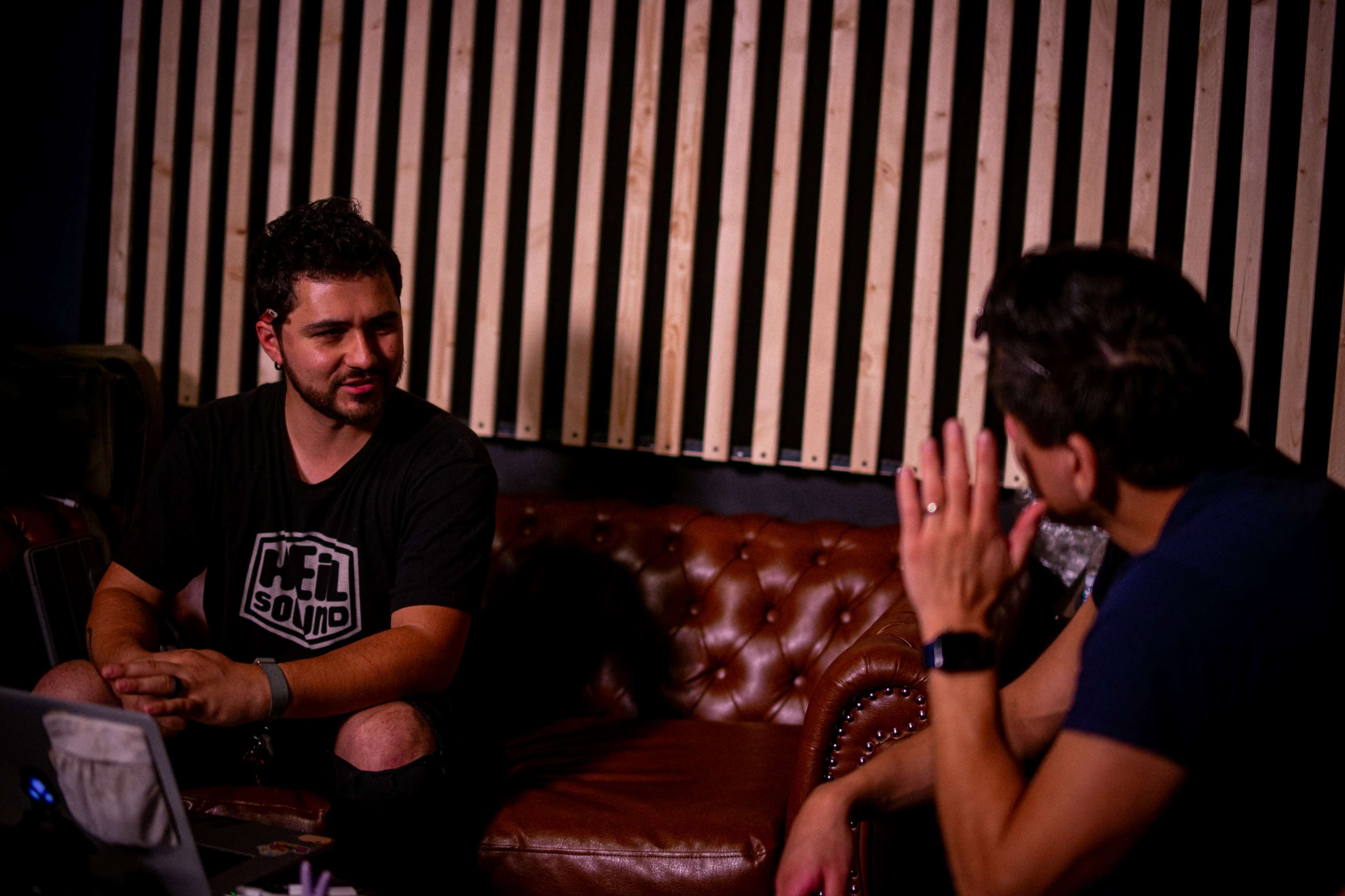 Two men are seated on a leather couch in a room with wooden paneling, engaged in a conversation.