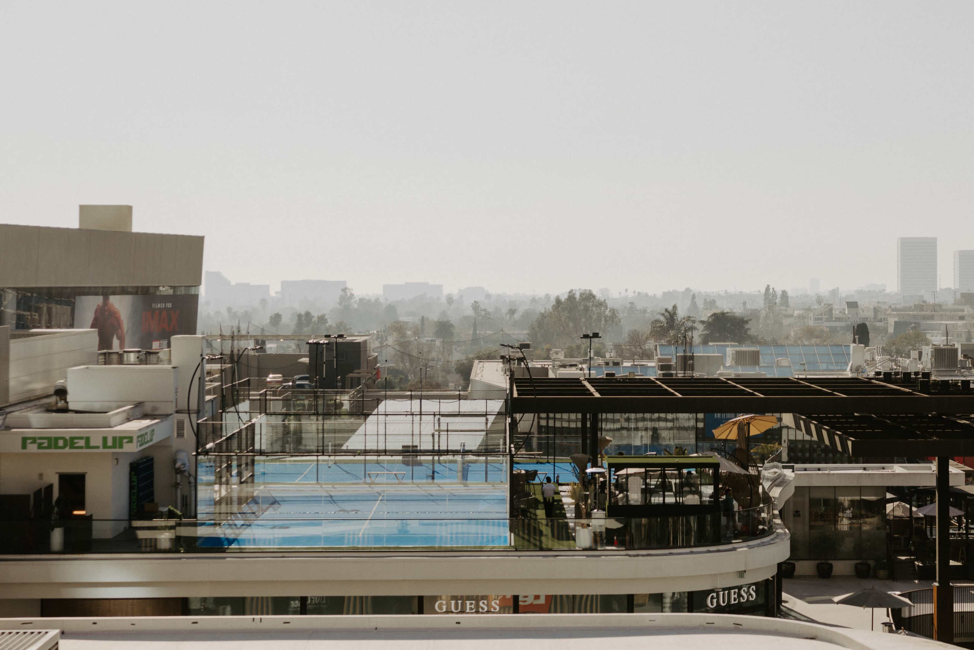 The image shows a rooftop view featuring a blue padel court and several buildings in a city, with a hazy skyline in the background.