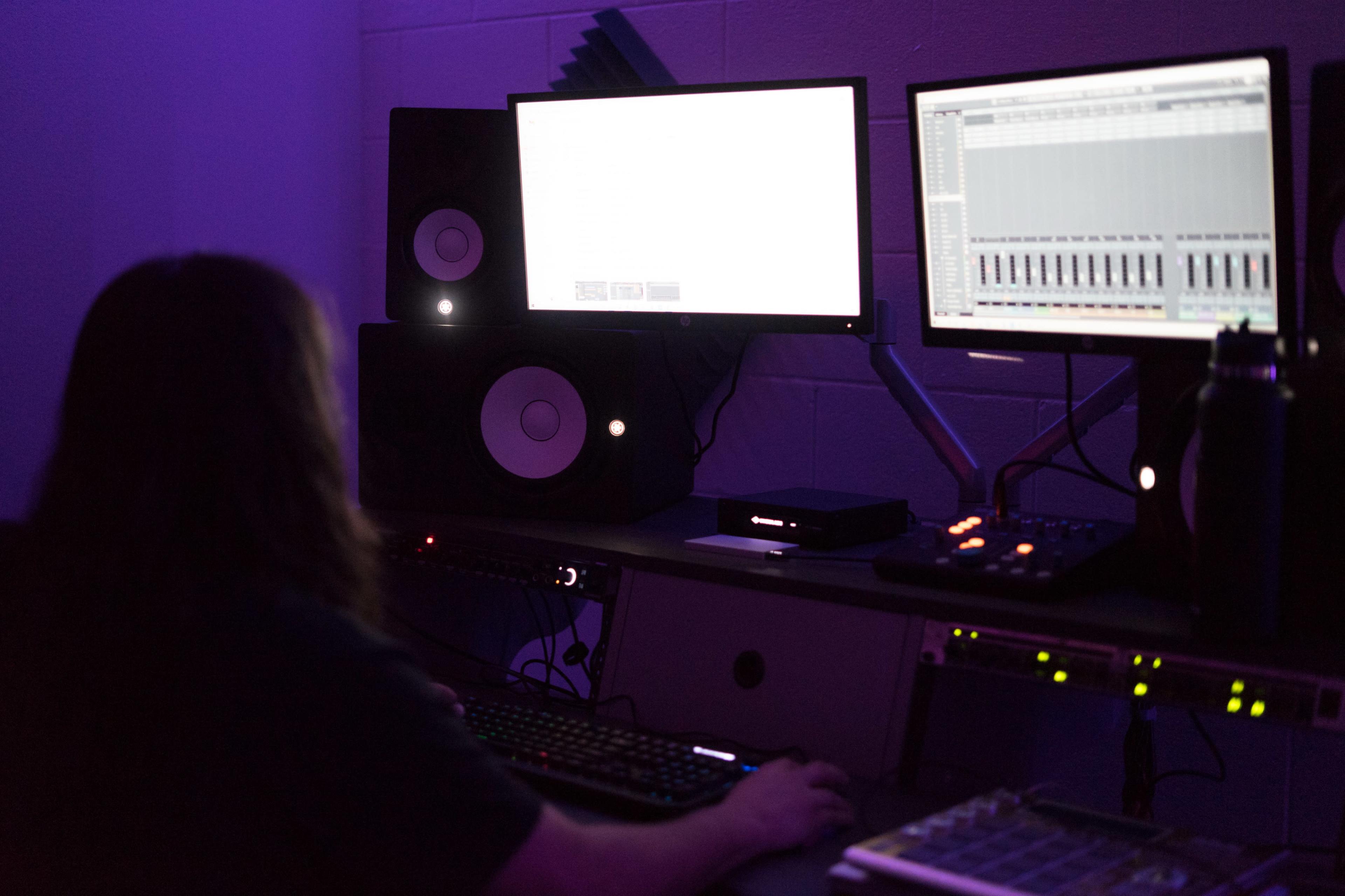 A person with long hair sits at a desk in a dimly lit room, focused on two computer monitors surrounded by audio equipment.