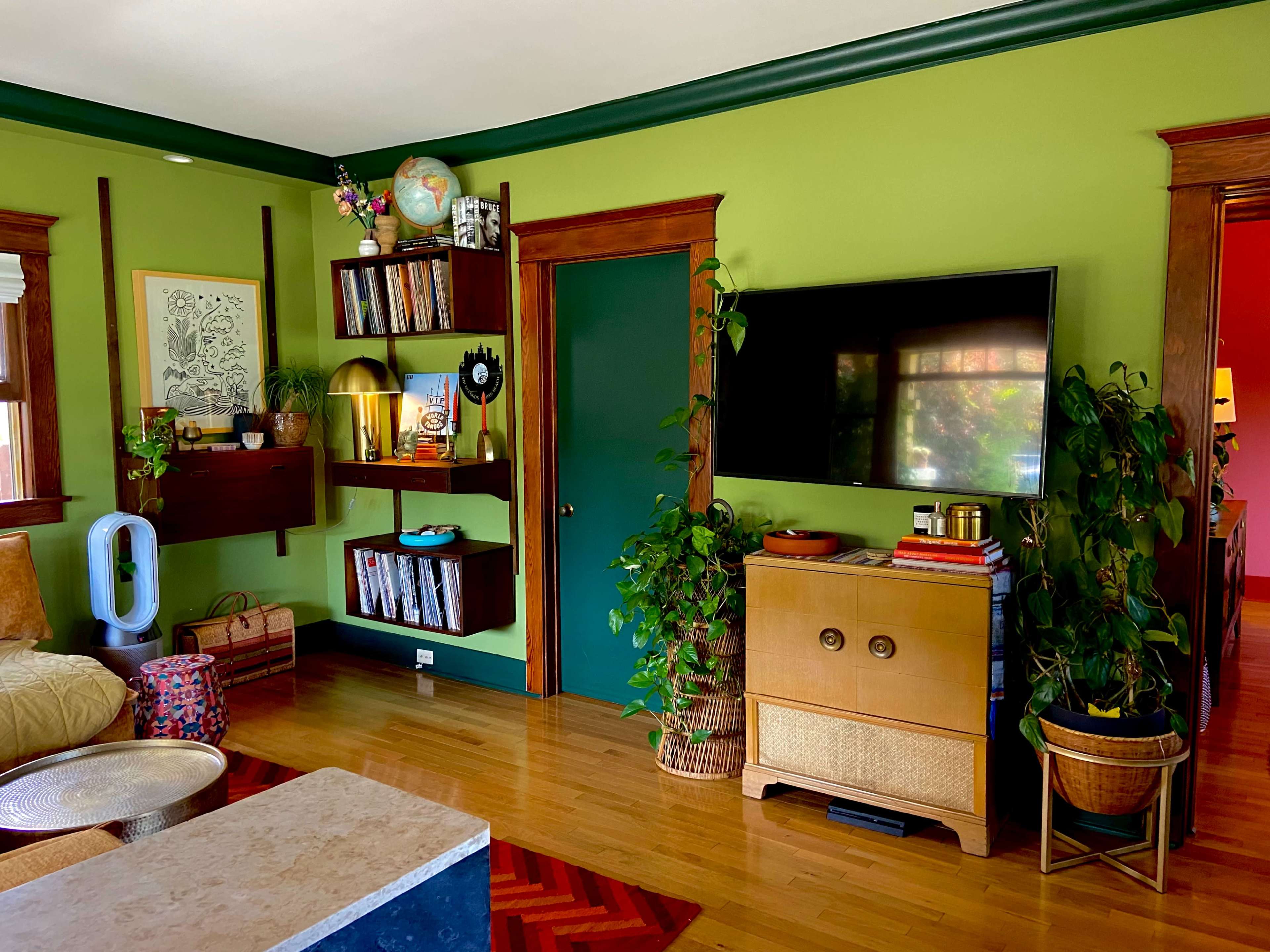 The image shows a living room with green walls, a television mounted on a wall, wooden furniture, shelves filled with books and decorative items, and several potted plants.