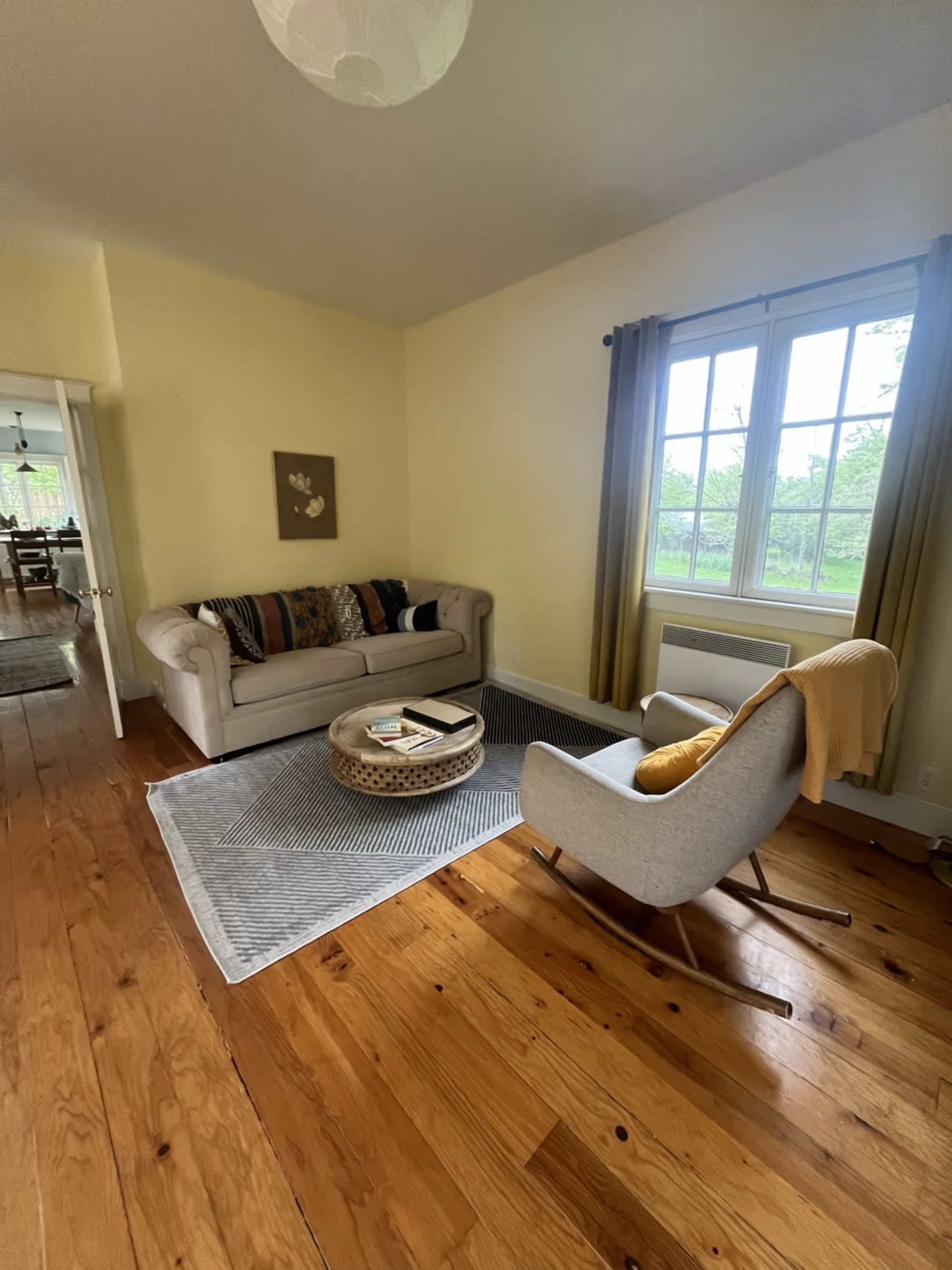The image shows a cozy living room with a light-colored couch, a round coffee table on a patterned rug, and a comfortable armchair beside a window.