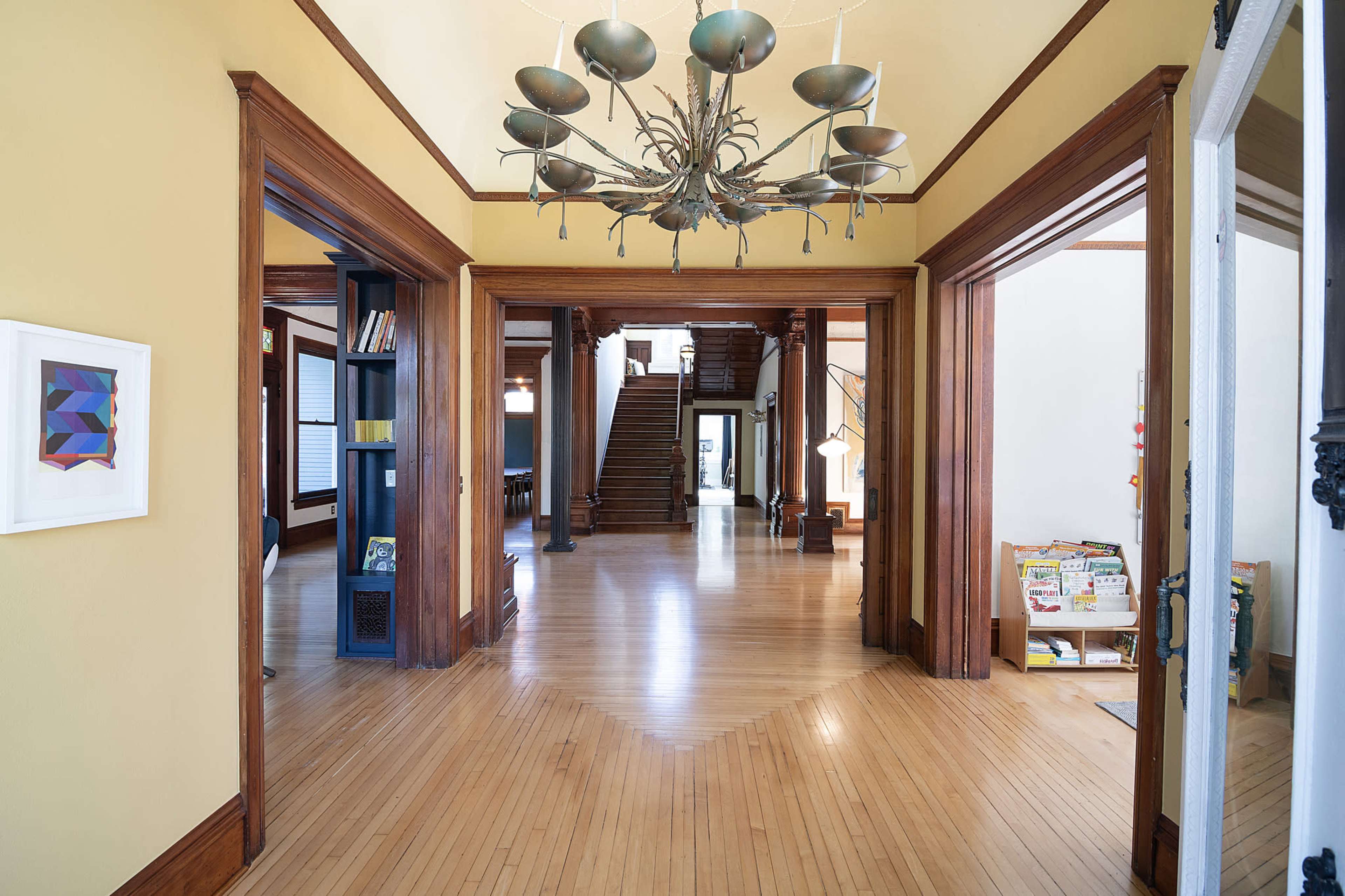 The image shows a spacious hallway with wooden flooring, intricate door frames, and a large chandelier hanging from the ceiling, leading to a staircase at the far end.
