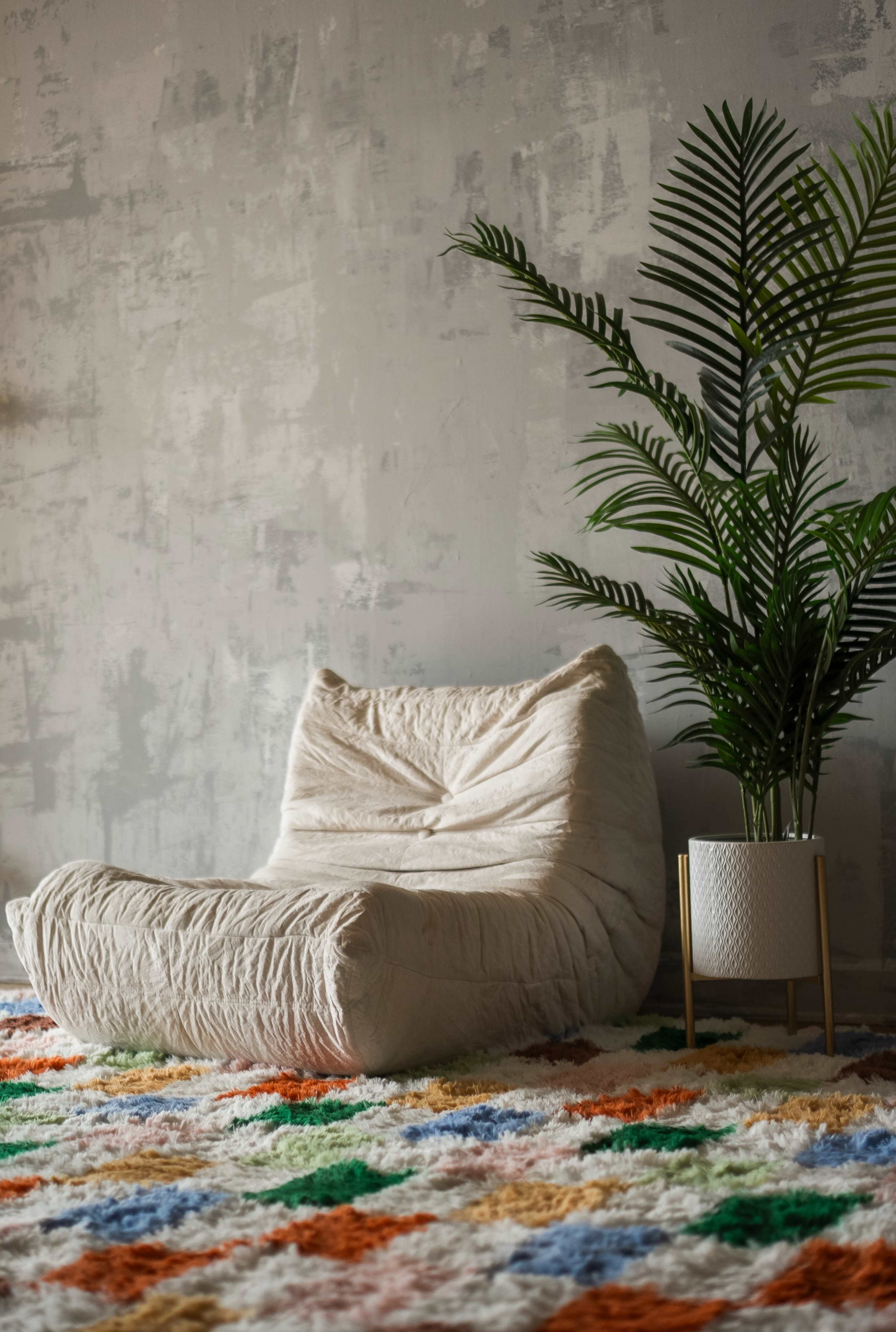 A comfortable, cream-colored chair is positioned on a colorful, patterned rug next to a potted plant in a decorative planter against a textured wall.