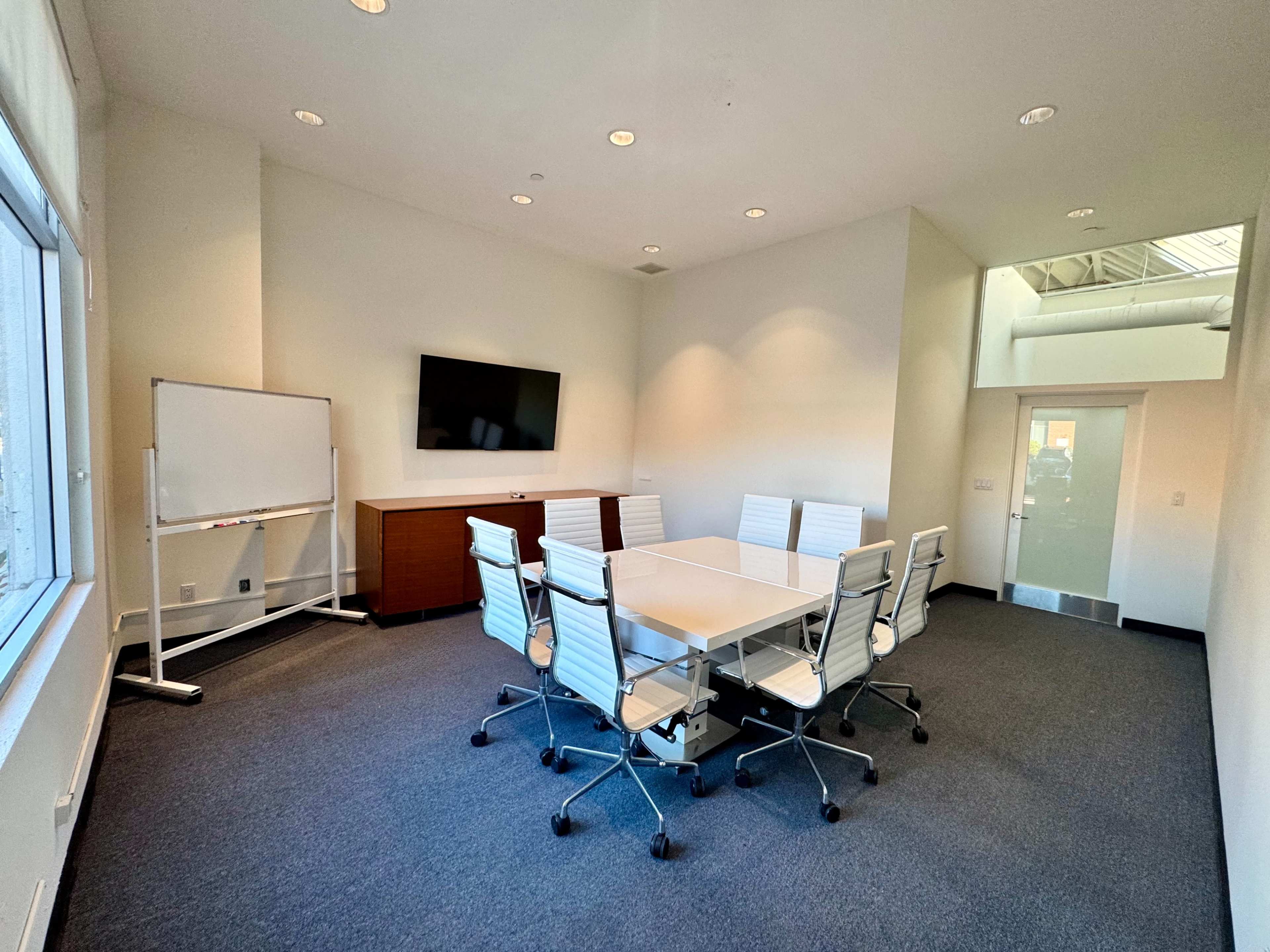 The image shows a modern conference room with a rectangular table, eight white chairs, a television on the wall, a whiteboard on a stand, and large windows letting in natural light.