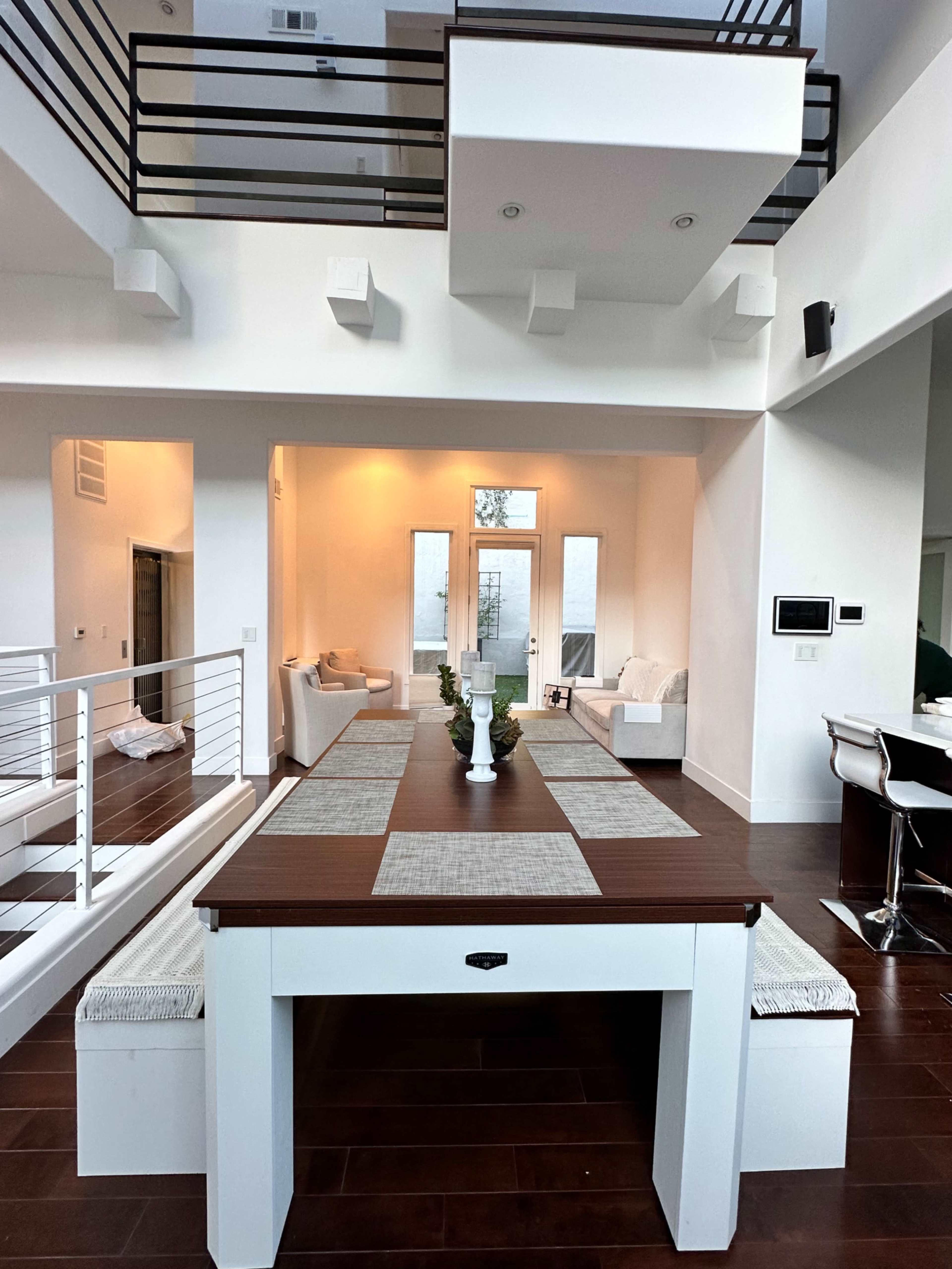 A modern, open-concept living space with a large dining table at the center, white sofas in the background, and large windows allowing natural light to enter.