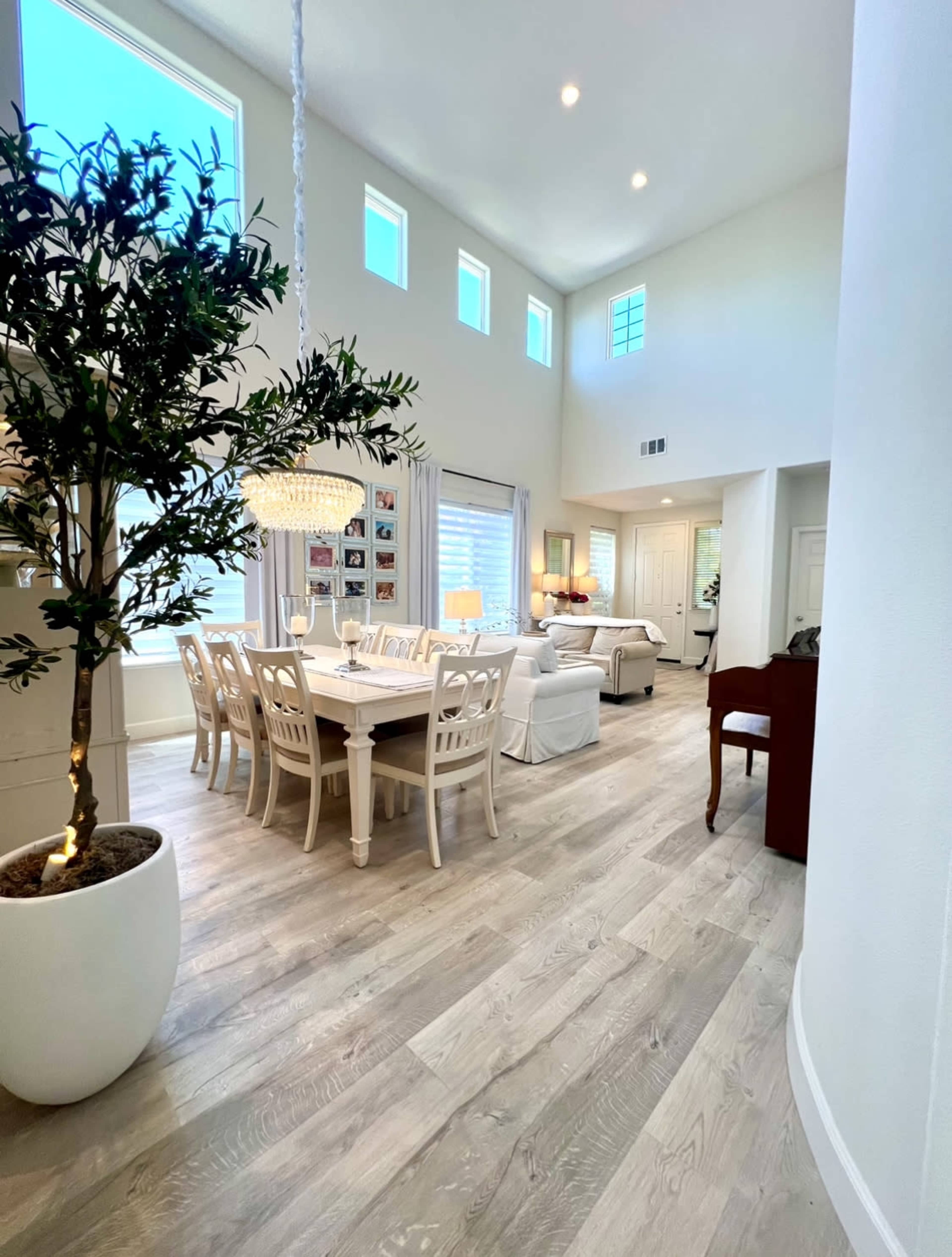A spacious, well-lit living and dining area with a wooden table surrounded by chairs, a chandelier overhead, and a large window allowing natural light in.