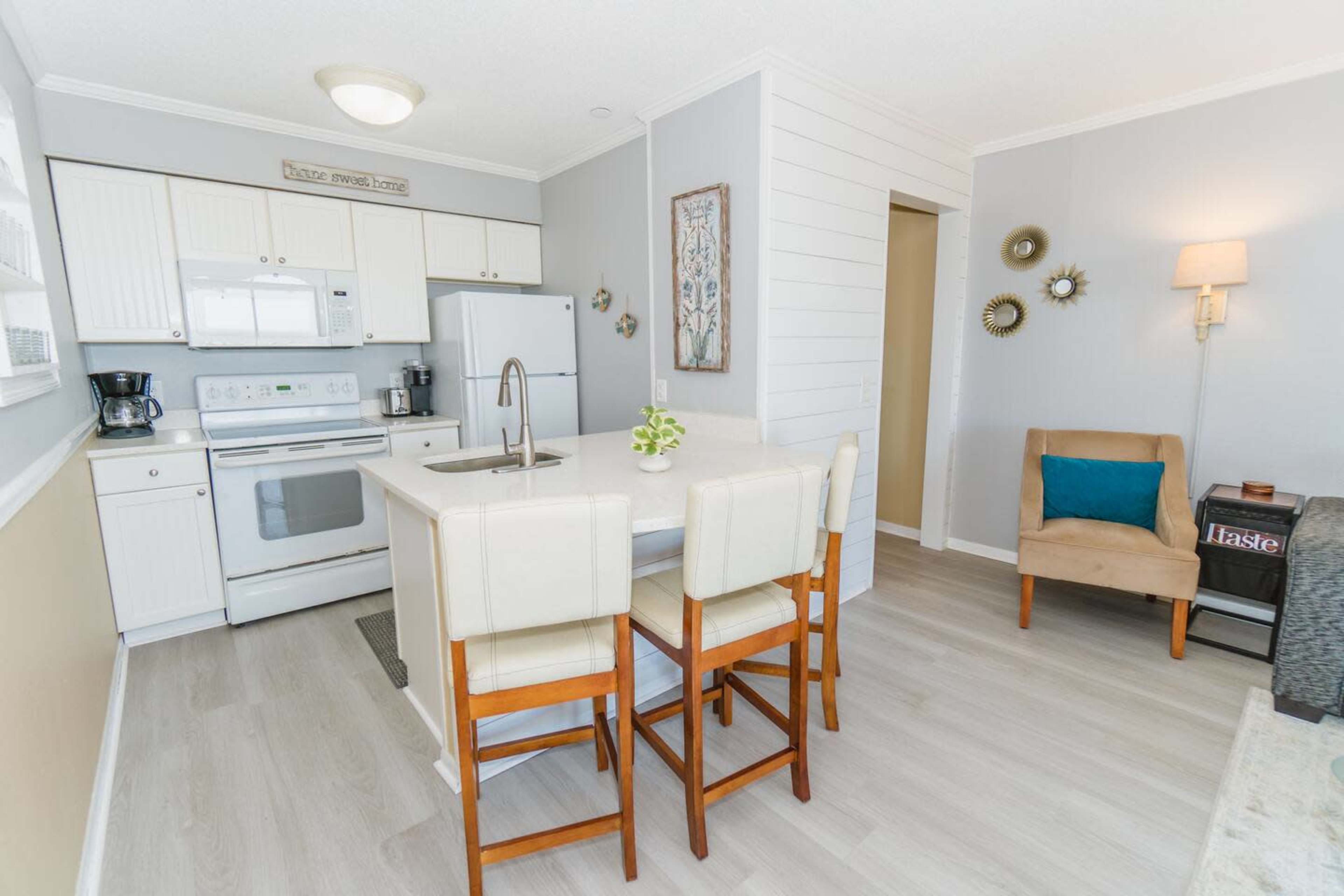 The image shows a modern kitchen with white cabinetry, a light gray accent wall, and a small dining area featuring four barstools at a counter.