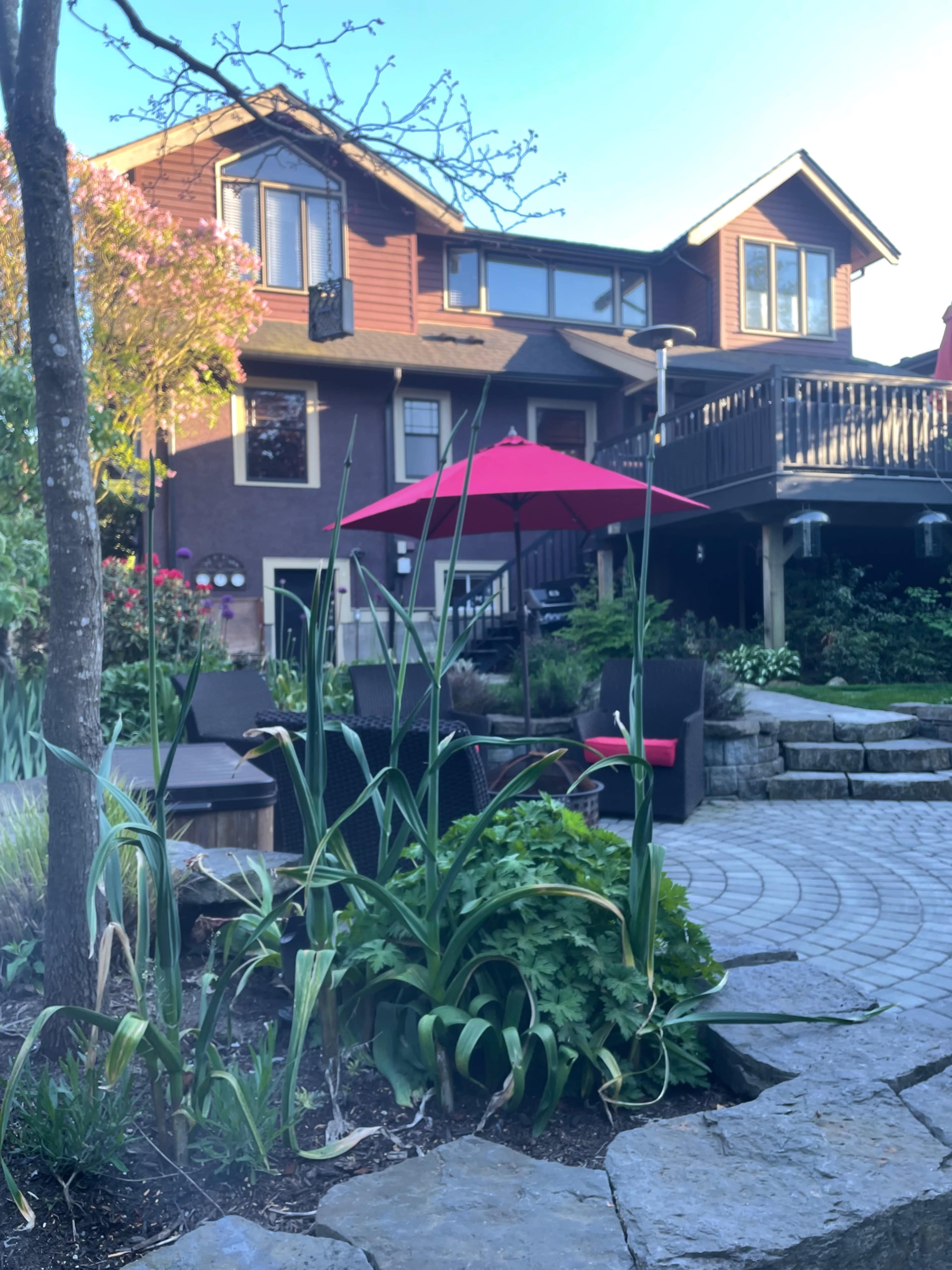 A patio area with outdoor furniture and a red umbrella is situated in front of a two-story brown house, surrounded by landscaped plants and stone pathways.