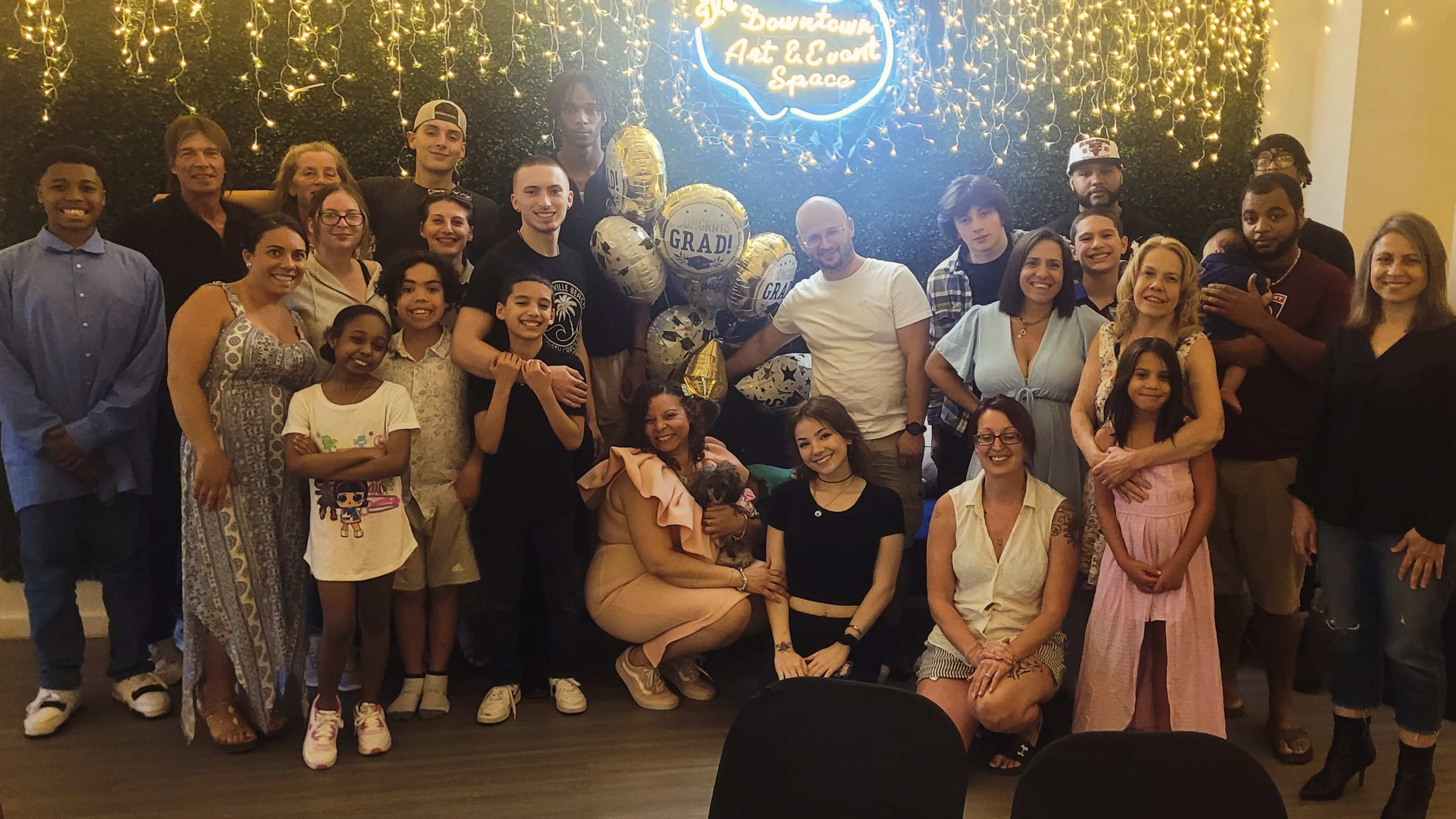 A large group of people poses together in a festive setting decorated with a "GRAD" balloon and twinkling lights.