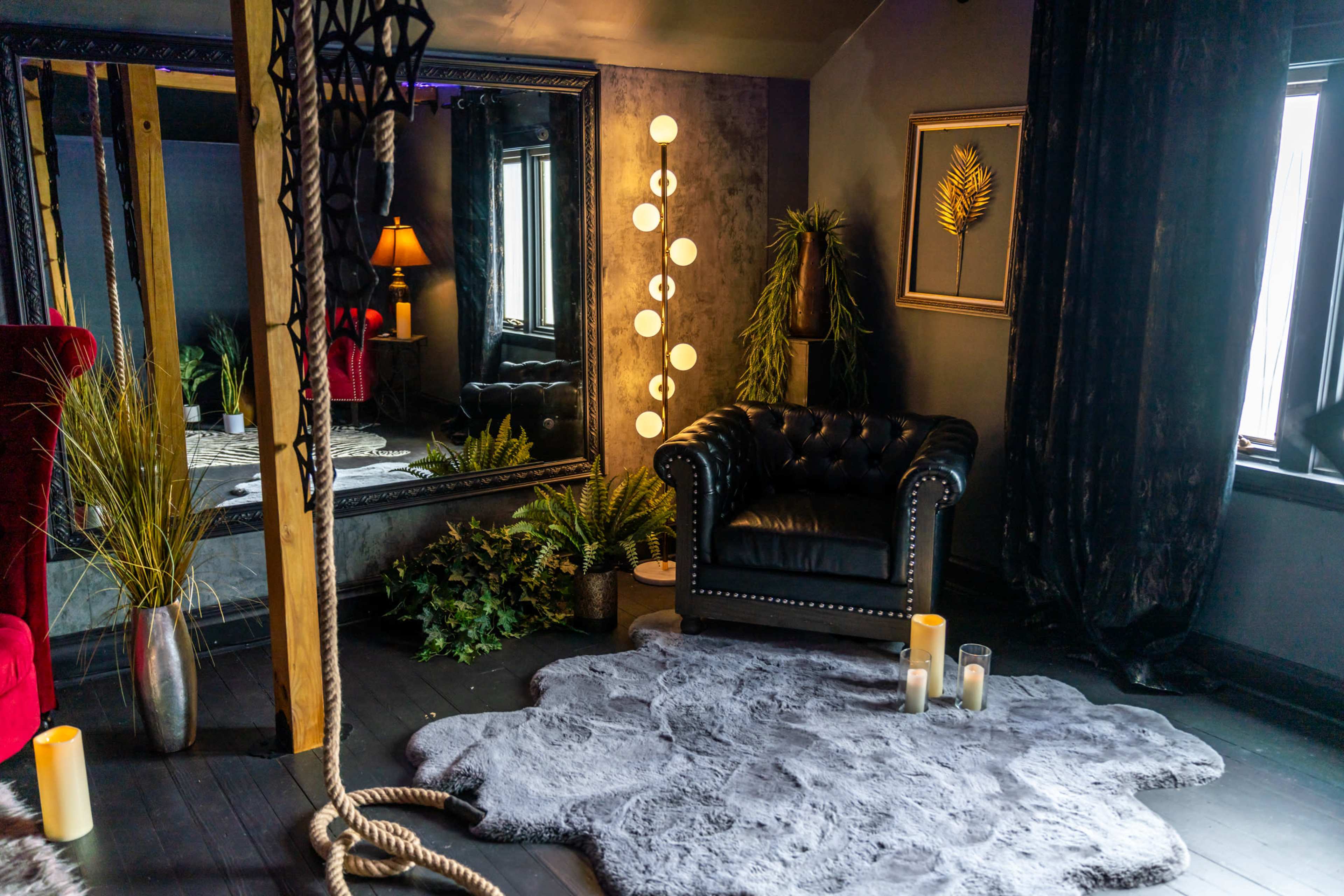 The scene features a dark room with a leather armchair, a large mirror, decorative plants, and soft lighting from floor lamps and candles.