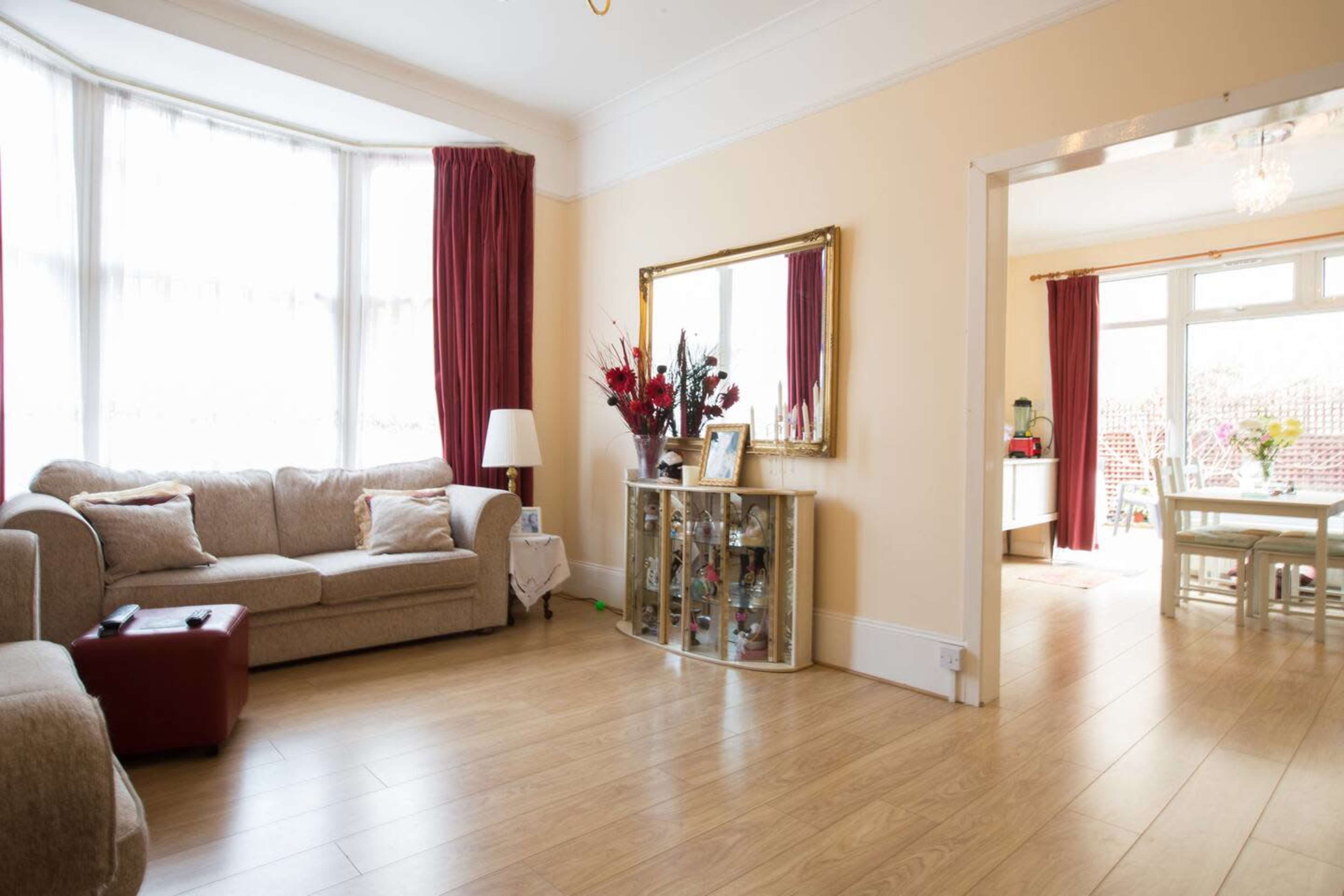 The image shows a bright living room with a beige sofa, decorative mirror, and a view into a dining area with a table and chairs.