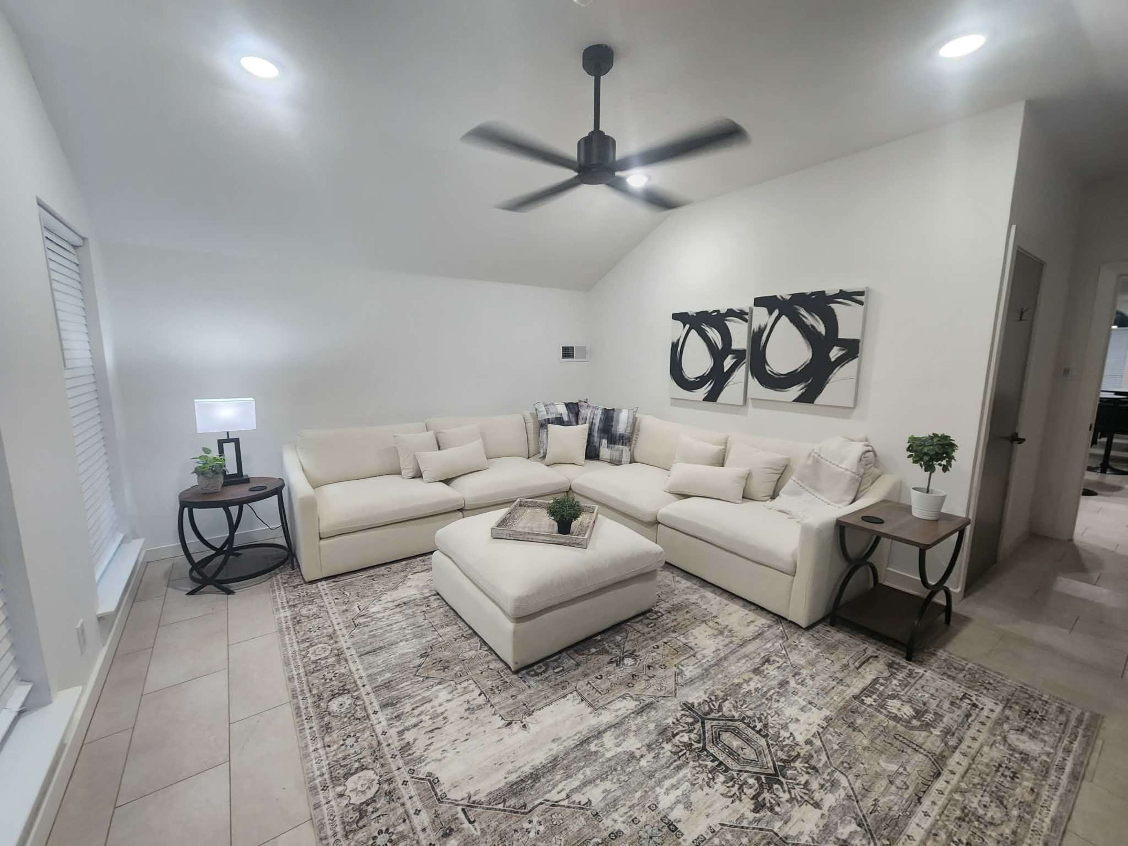 The image shows a modern living room featuring a large sectional sofa, an ottoman in the center, a small side table with a lamp, and two abstract wall paintings, all arranged on a patterned area rug.