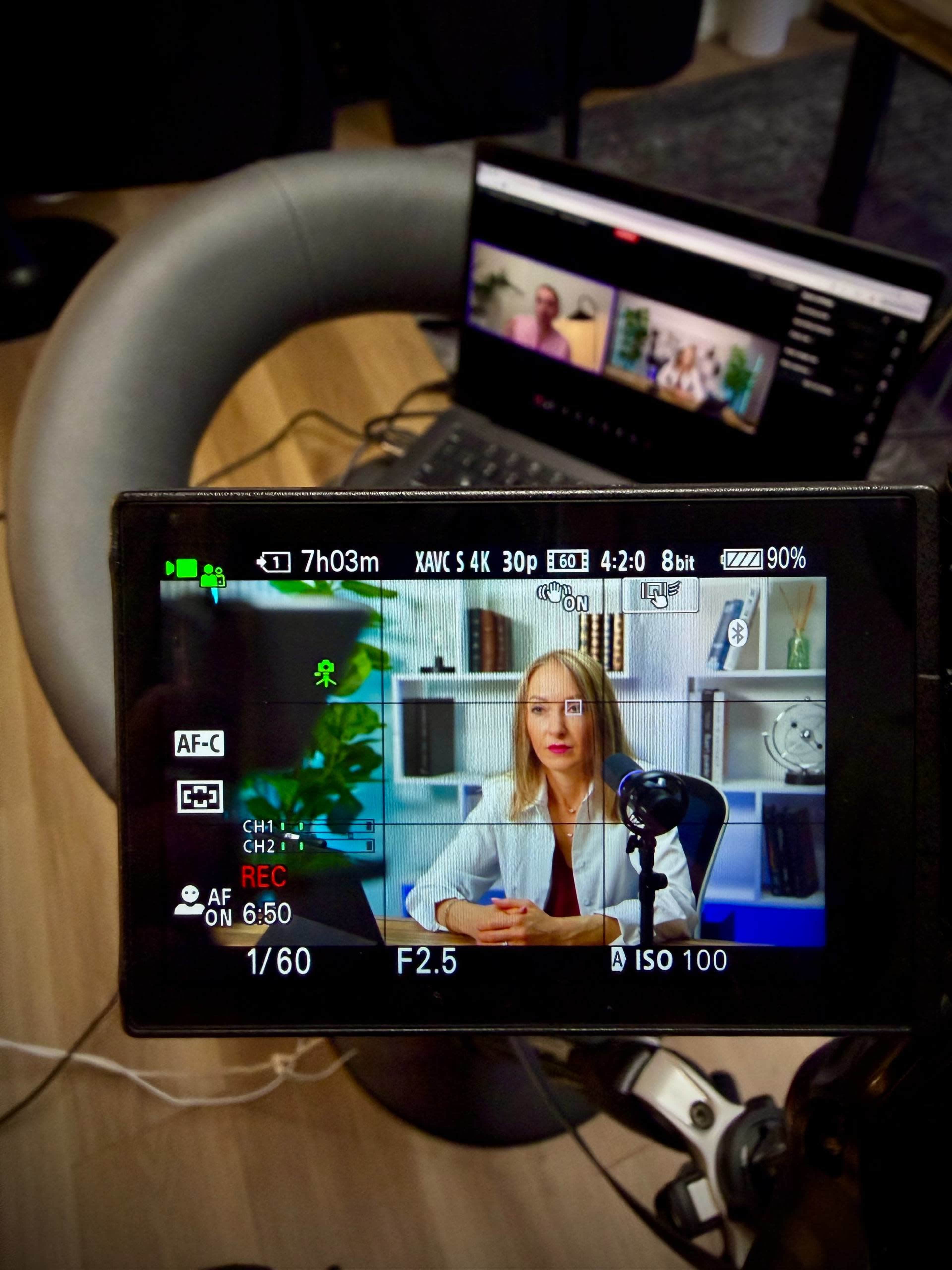 A camera viewfinder displays a woman speaking into a microphone, with a laptop showing video content in the background.
