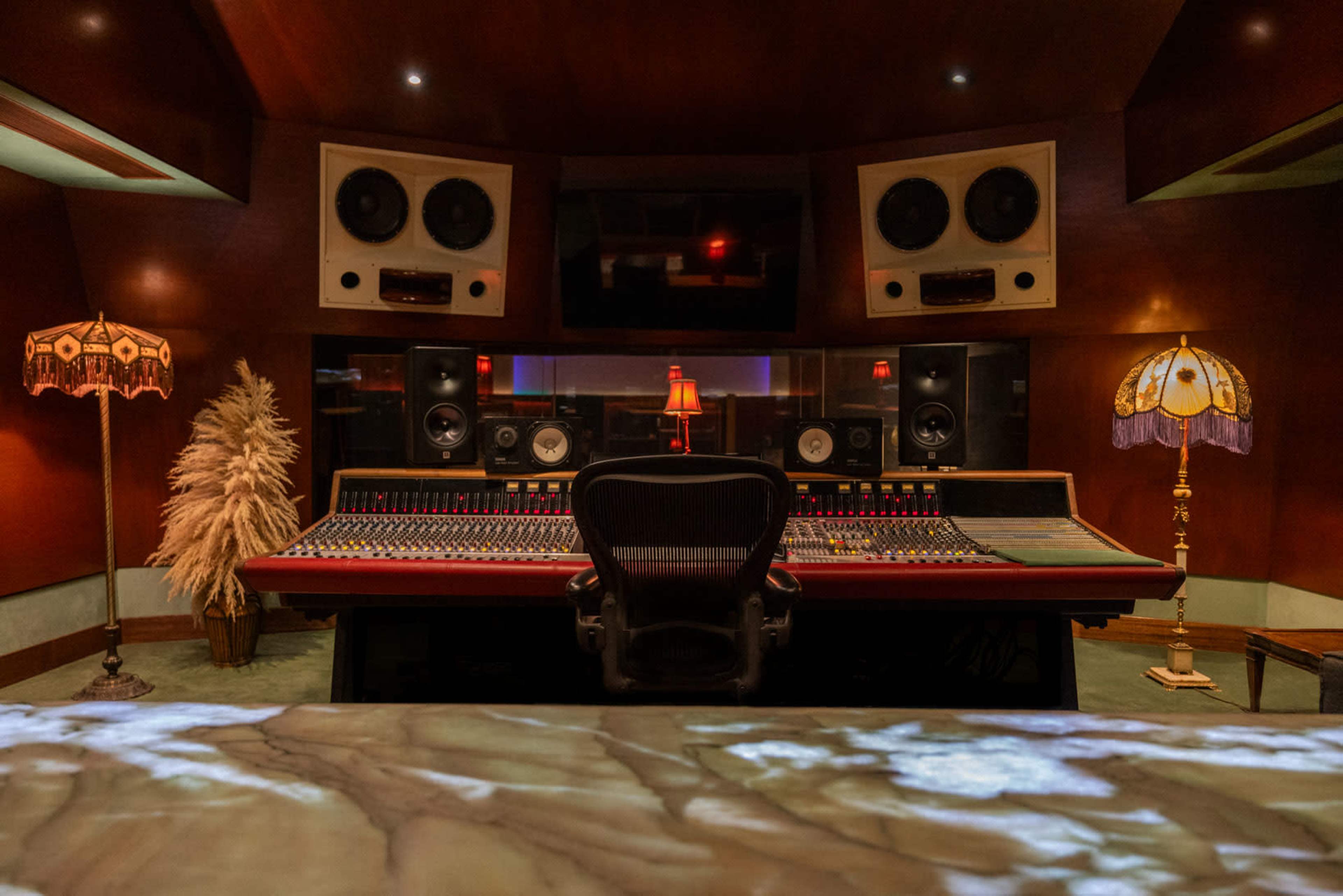 The image shows a dimly lit recording studio with a large mixing console, speakers mounted on the walls, and decorative lamps on either side.