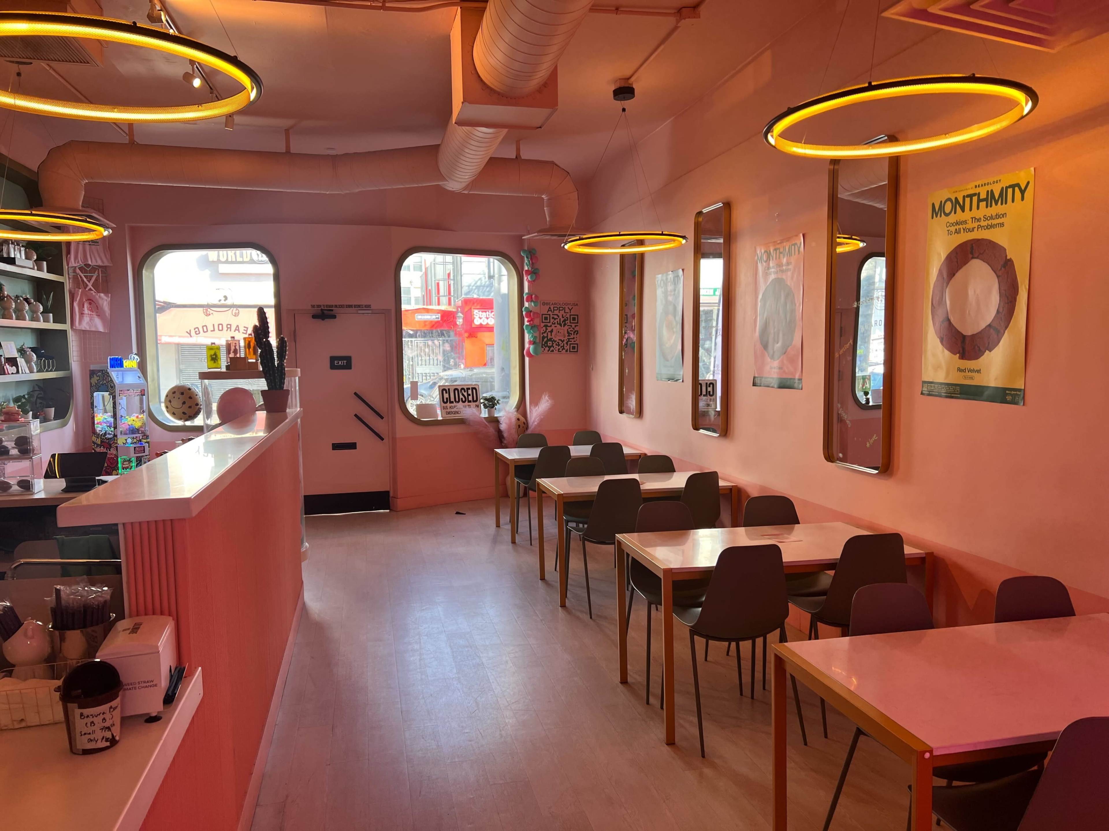 The interior of a café features pink walls, wooden tables, and circular lights, with a view of the street through large windows.