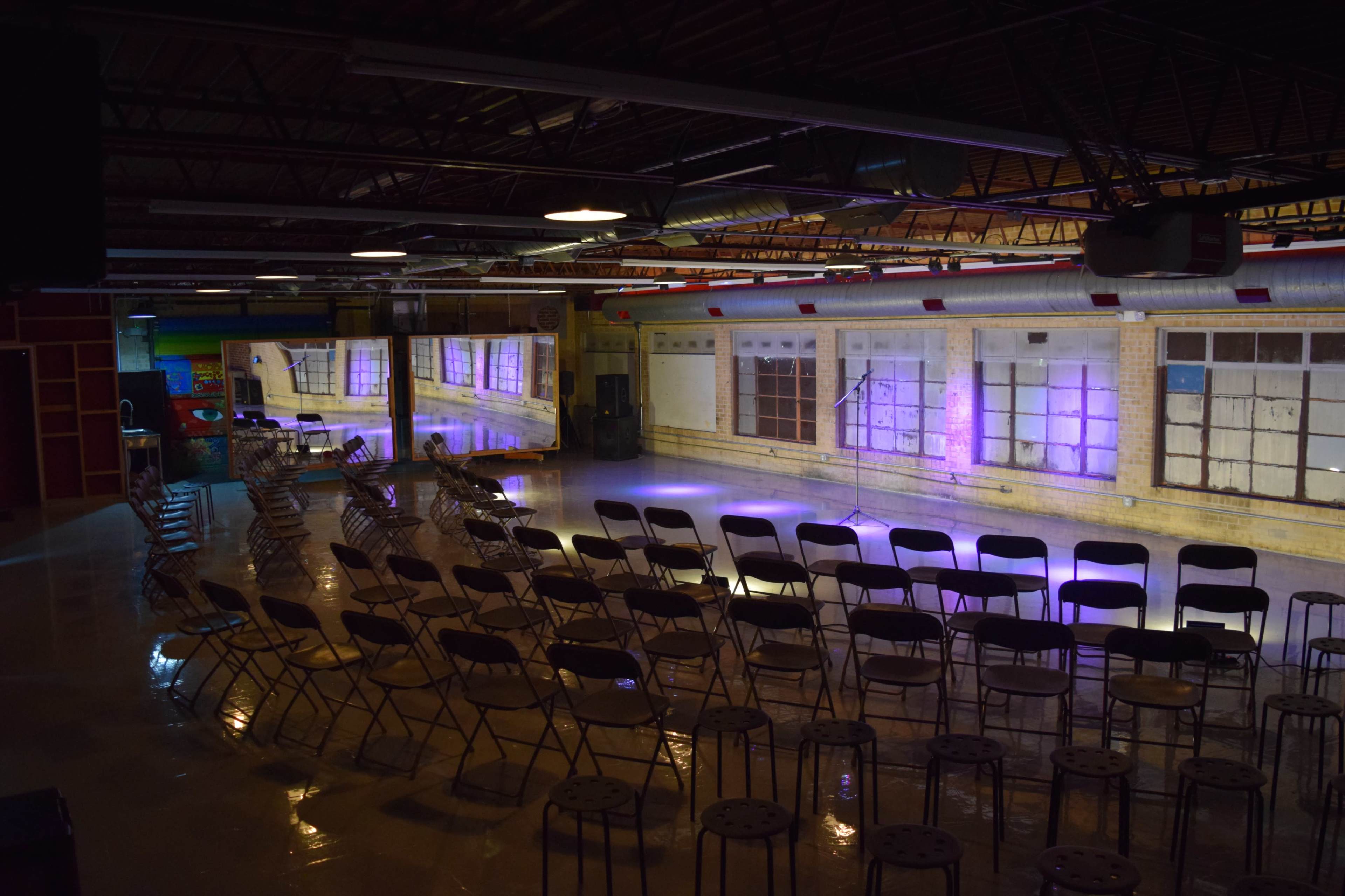 A spacious indoor venue set up with rows of chairs facing a stage, illuminated by soft purple lighting against a backdrop of large windows.