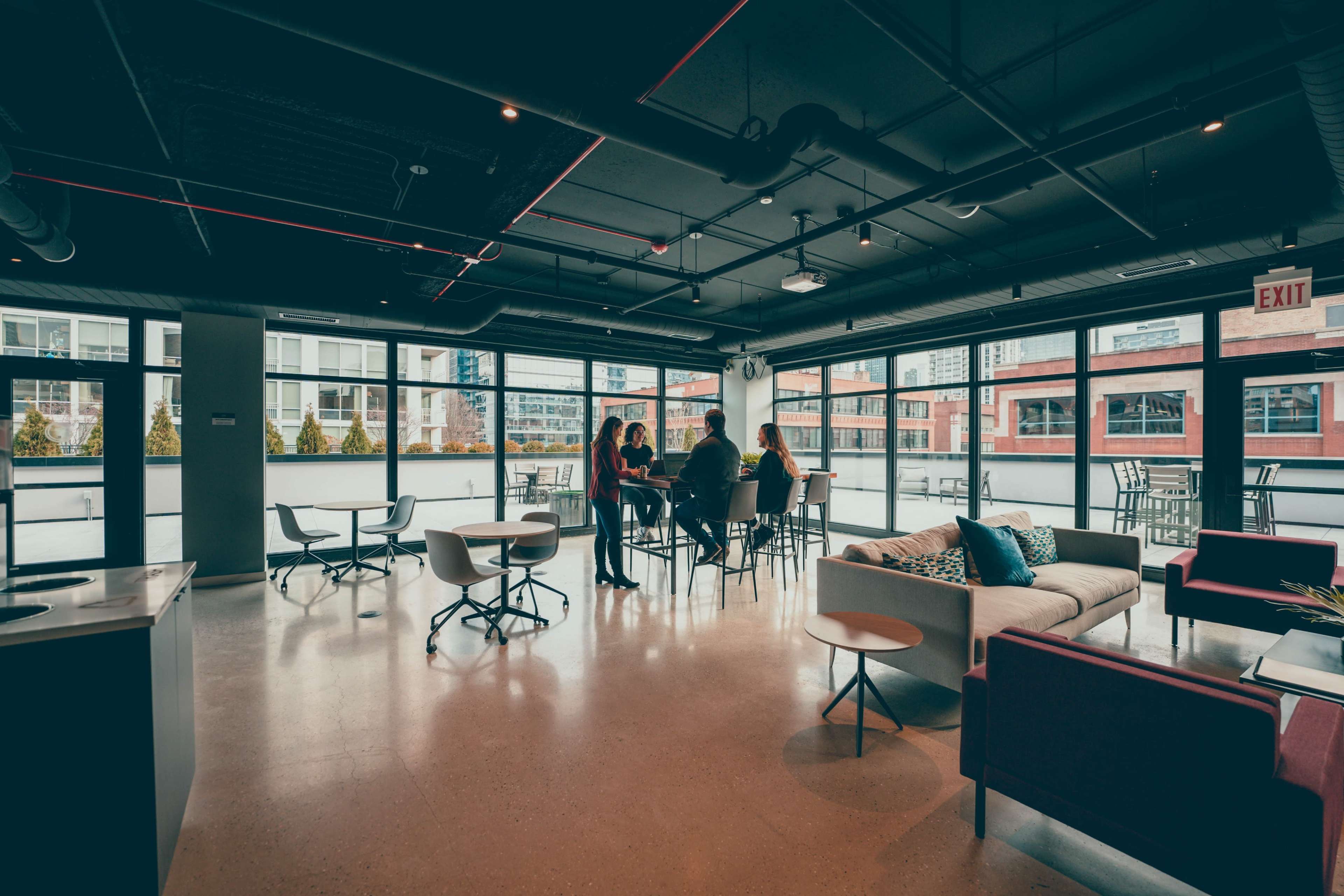A modern office space features a seating area with a couch, tables, and chairs, where four people are engaged in conversation near large windows overlooking a cityscape.