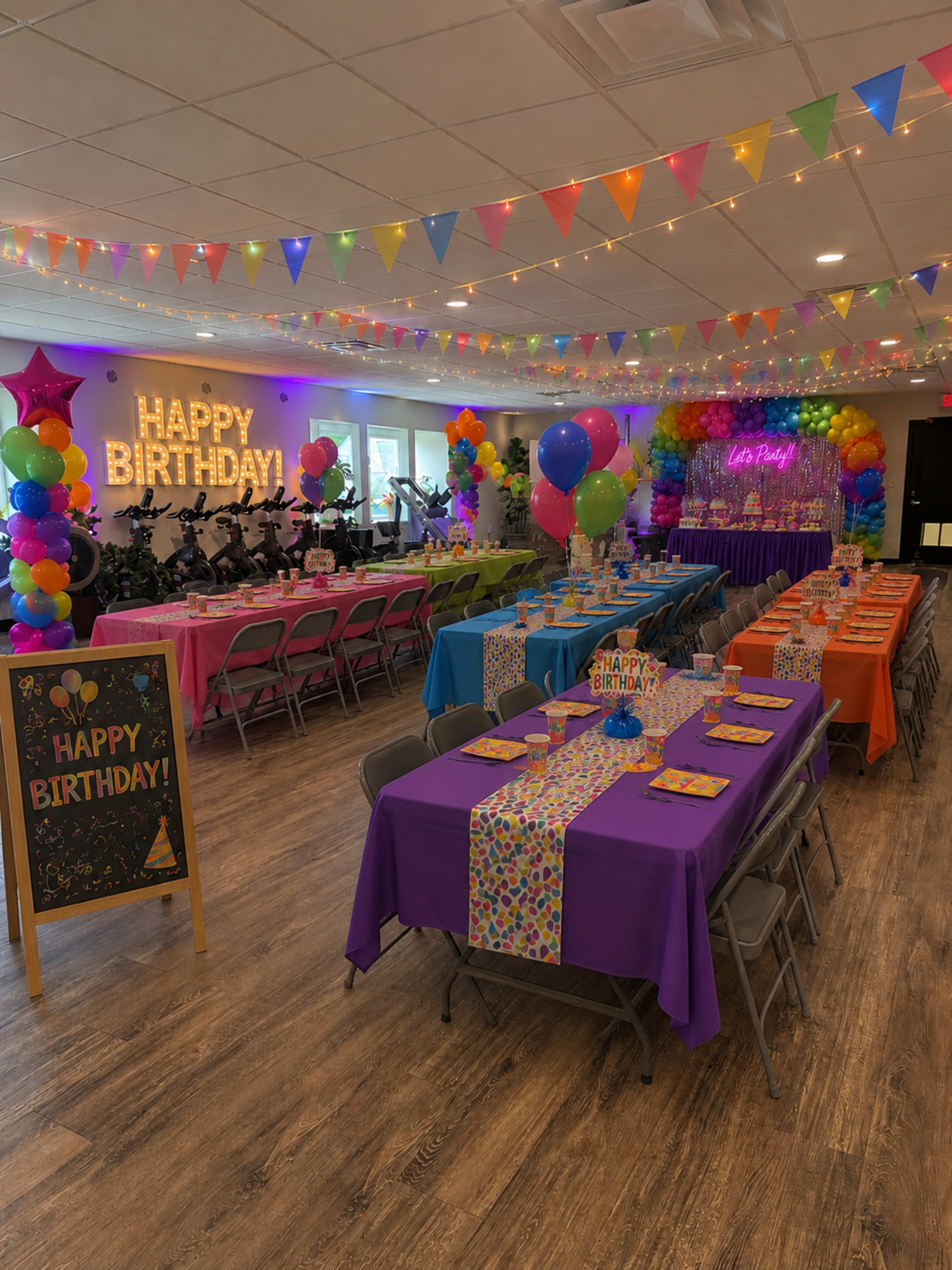 The image shows a brightly decorated party space with tables covered in colorful tablecloths, balloons, and a "Happy Birthday" sign illuminated against a wall.