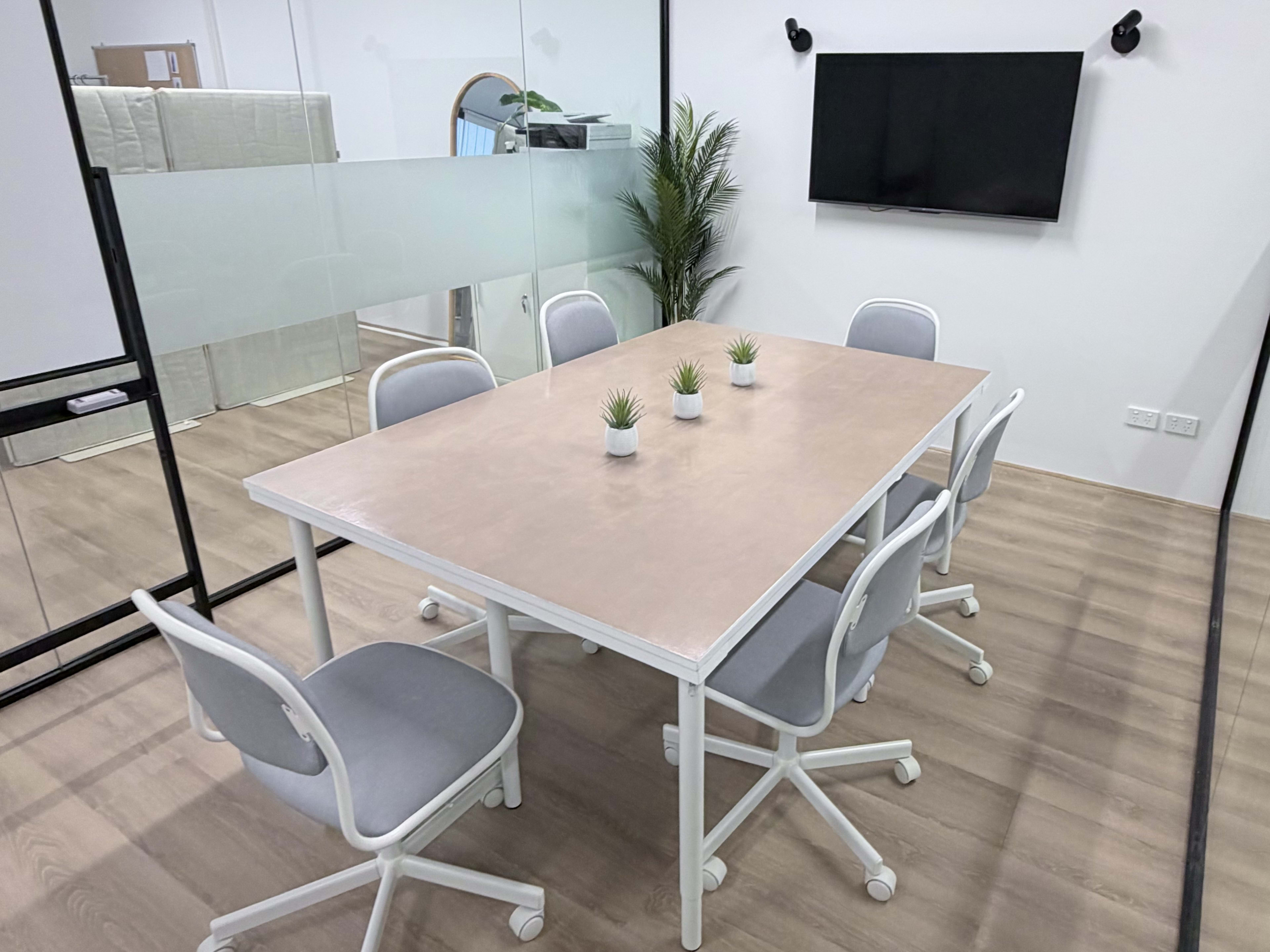 A modern conference room features a rectangular table surrounded by six chairs, with a television mounted on the wall and a potted plant in the corner.