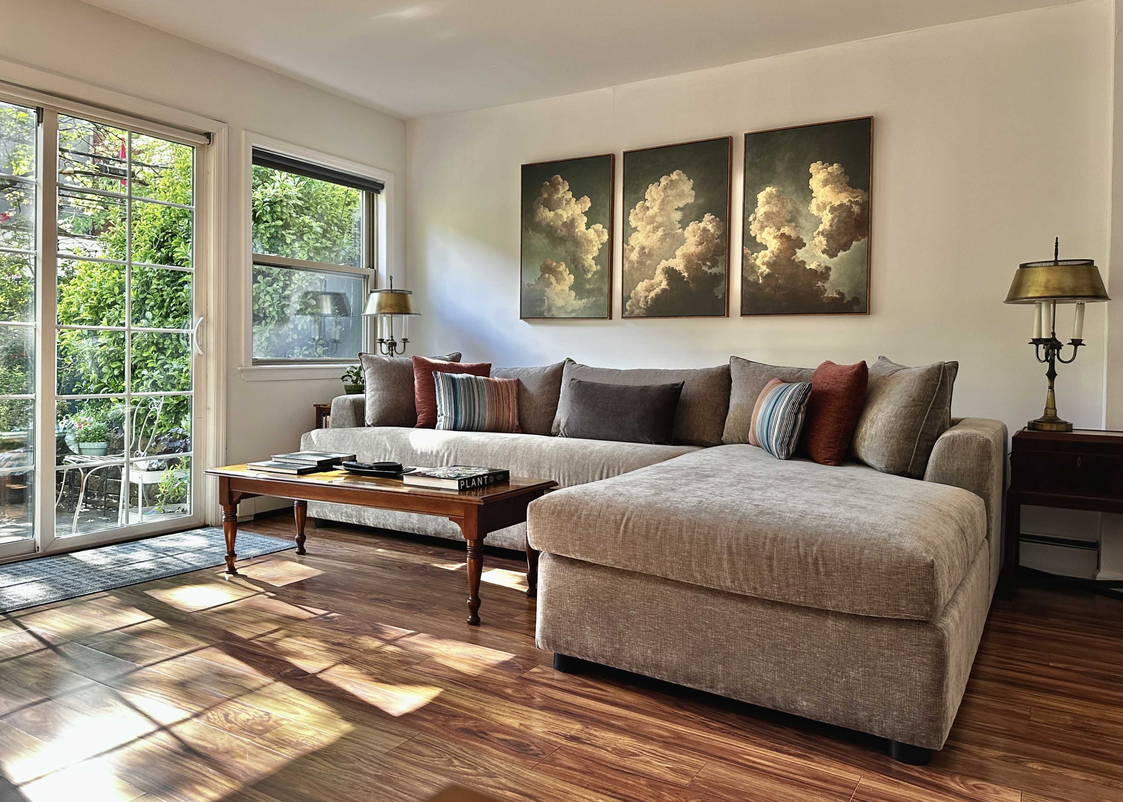 The image shows a cozy living room with a sectional sofa, a wooden coffee table, and three framed cloud paintings on the wall, illuminated by natural light from a window.