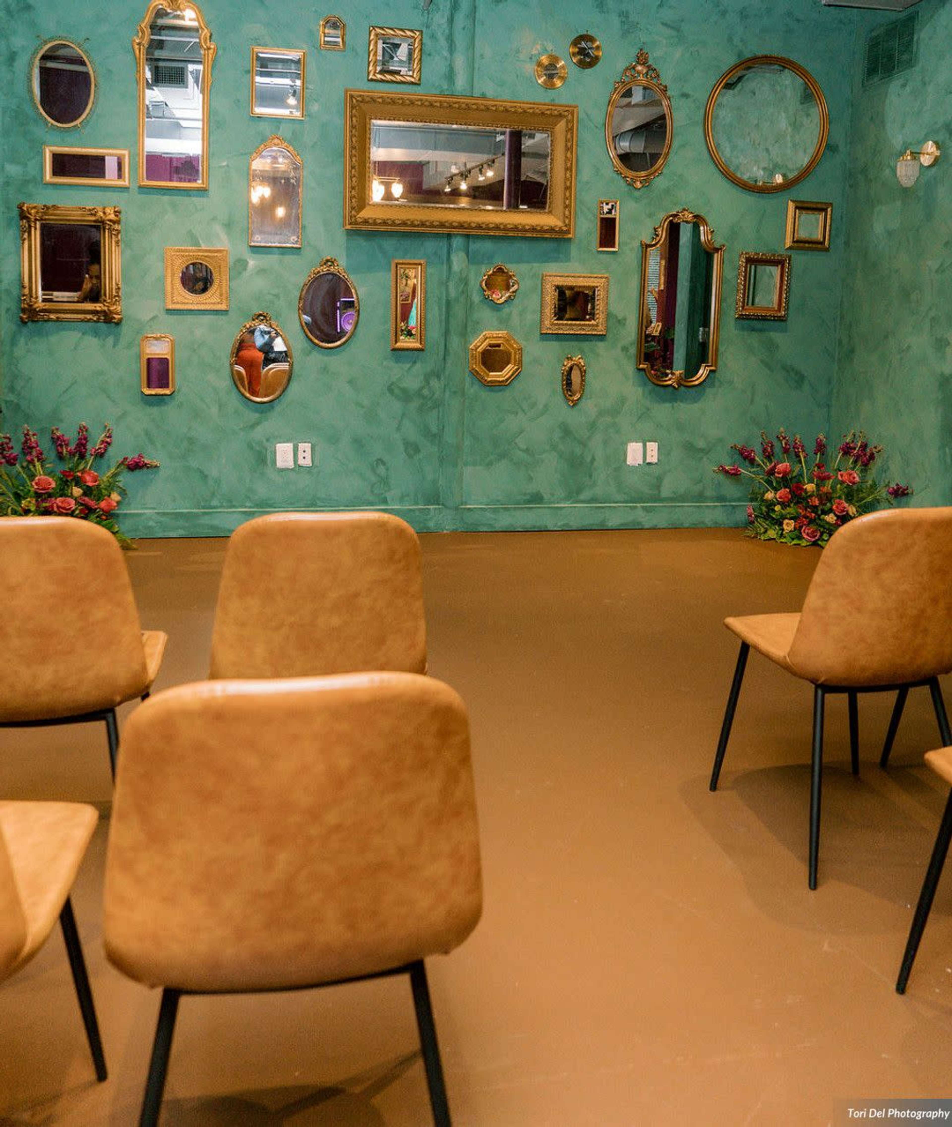 A room with a green textured wall featuring various mirrors in ornate frames and three empty brown chairs facing the mirrors.