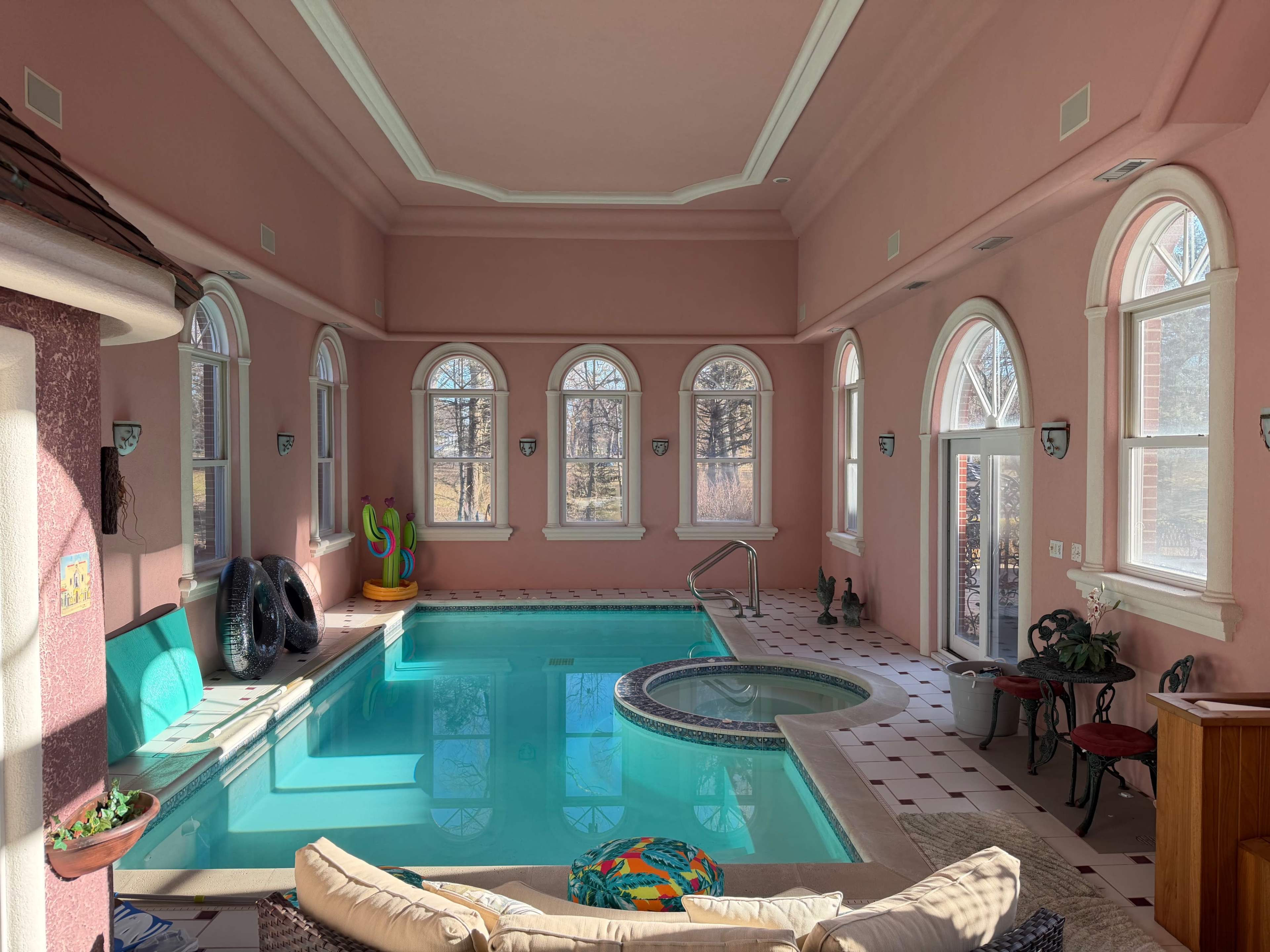 The image shows an indoor pool area with pink walls, large windows, and a small spa section next to the main pool.