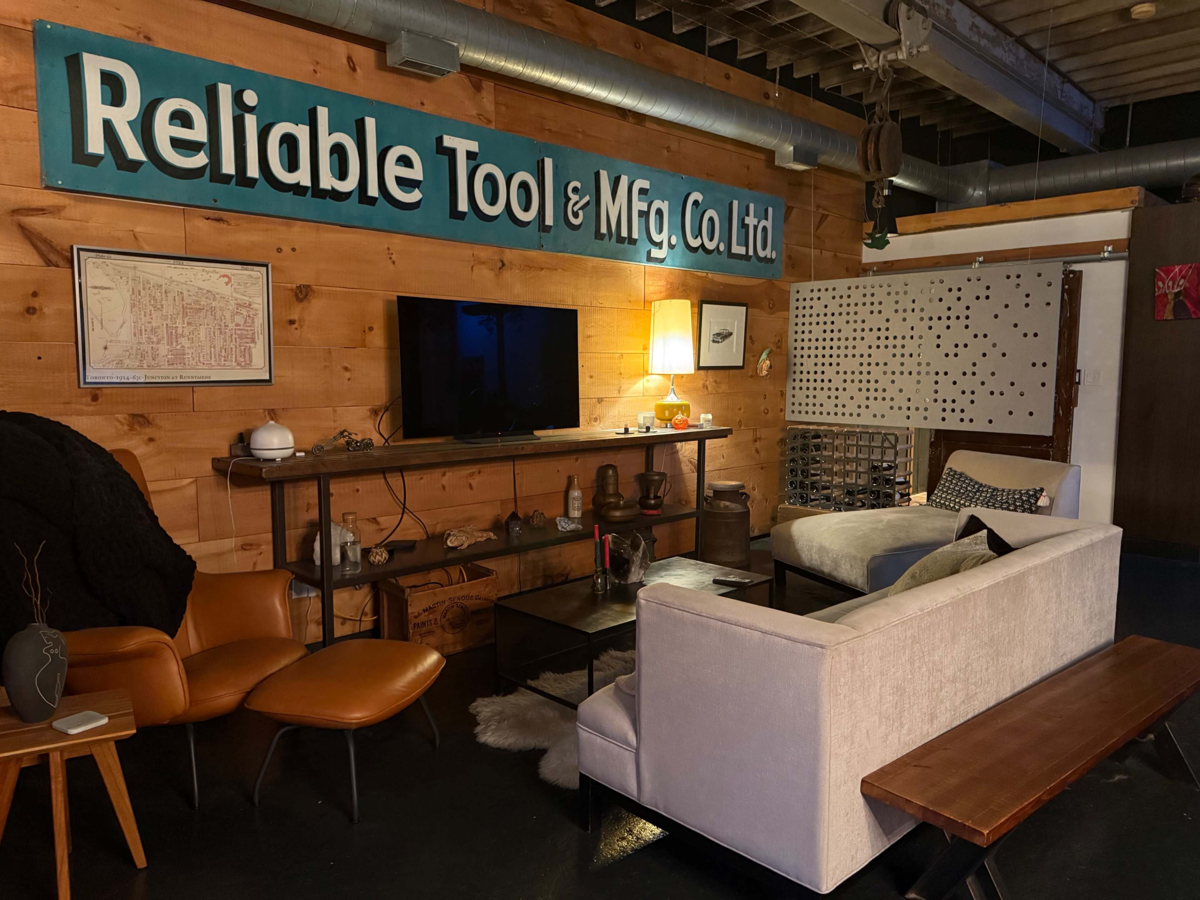 The scene shows a cozy living area with a wooden wall featuring the sign "Reliable Tool & Mfg. Co. Ltd., a television on a shelf, a couch, and a wooden bench.