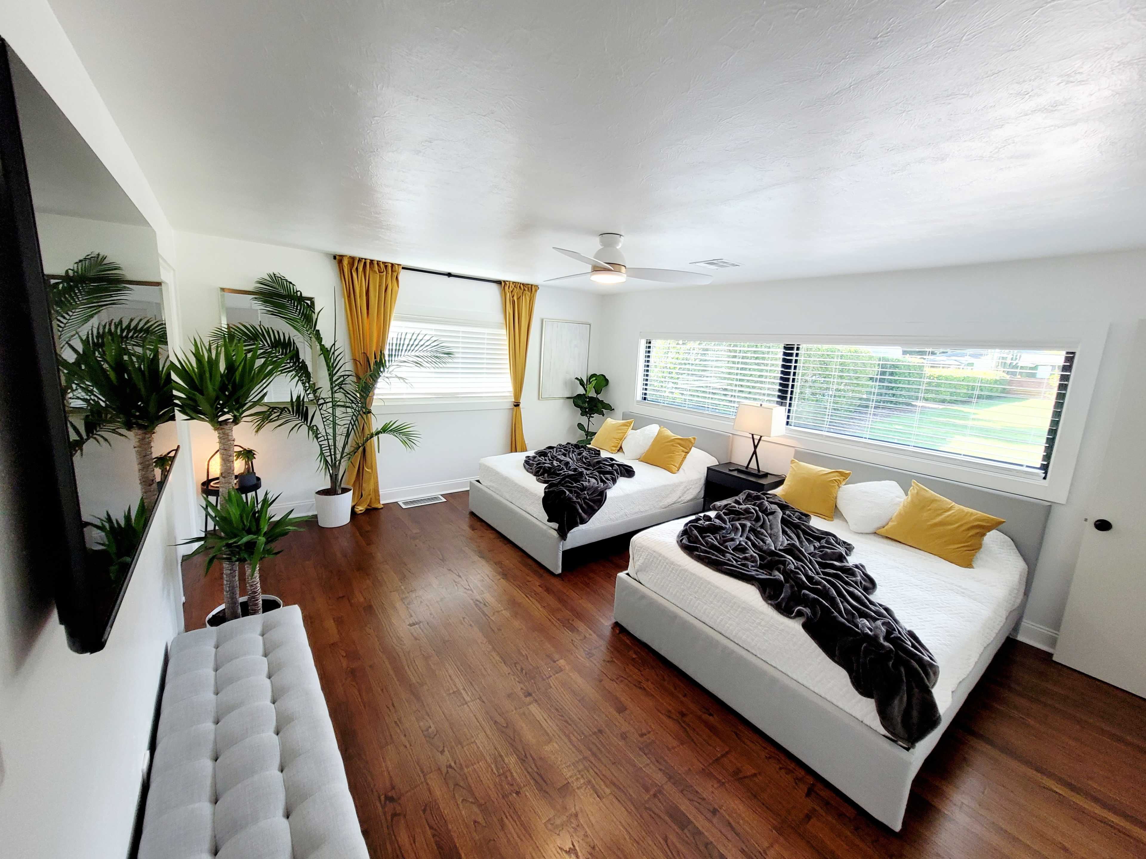 A bright, modern bedroom featuring two white beds, yellow accent pillows, and large windows with plants and a TV mounted on the wall.