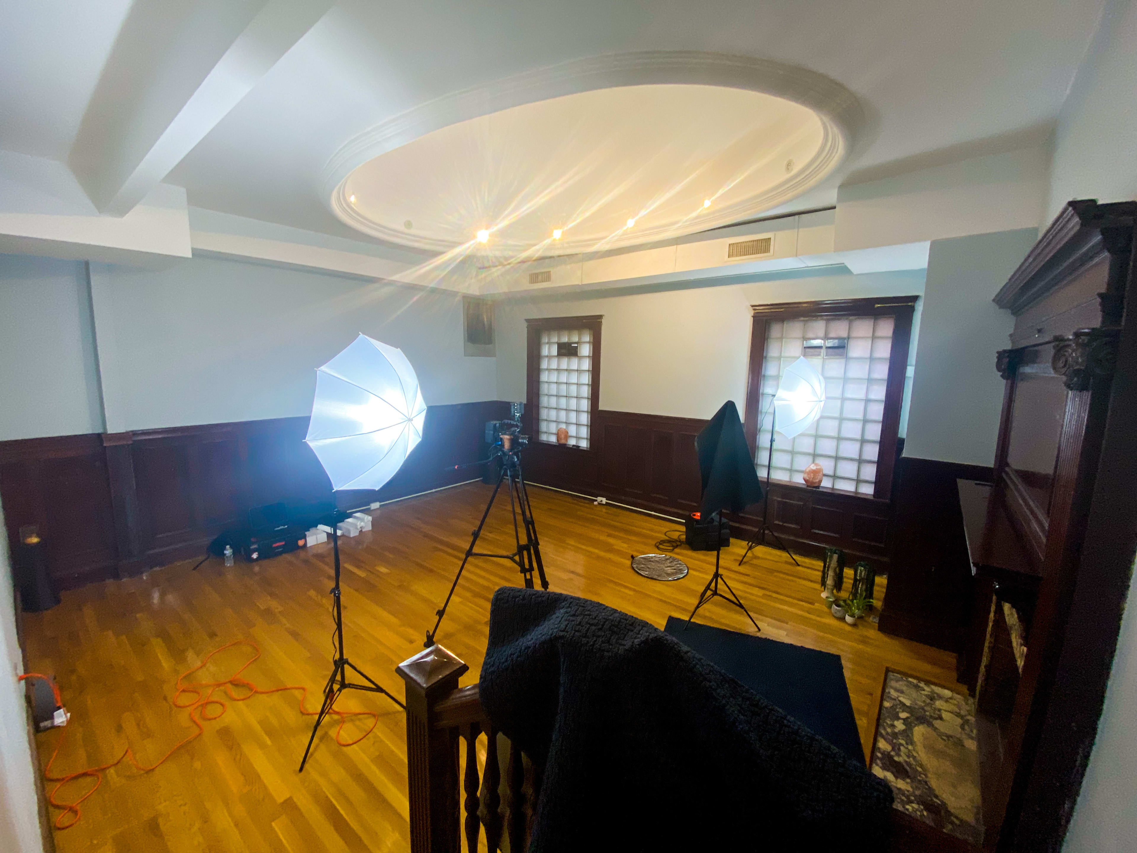 A vacant room with wooden floors, two windows, and photography equipment including softbox lights and a camera on a tripod.
