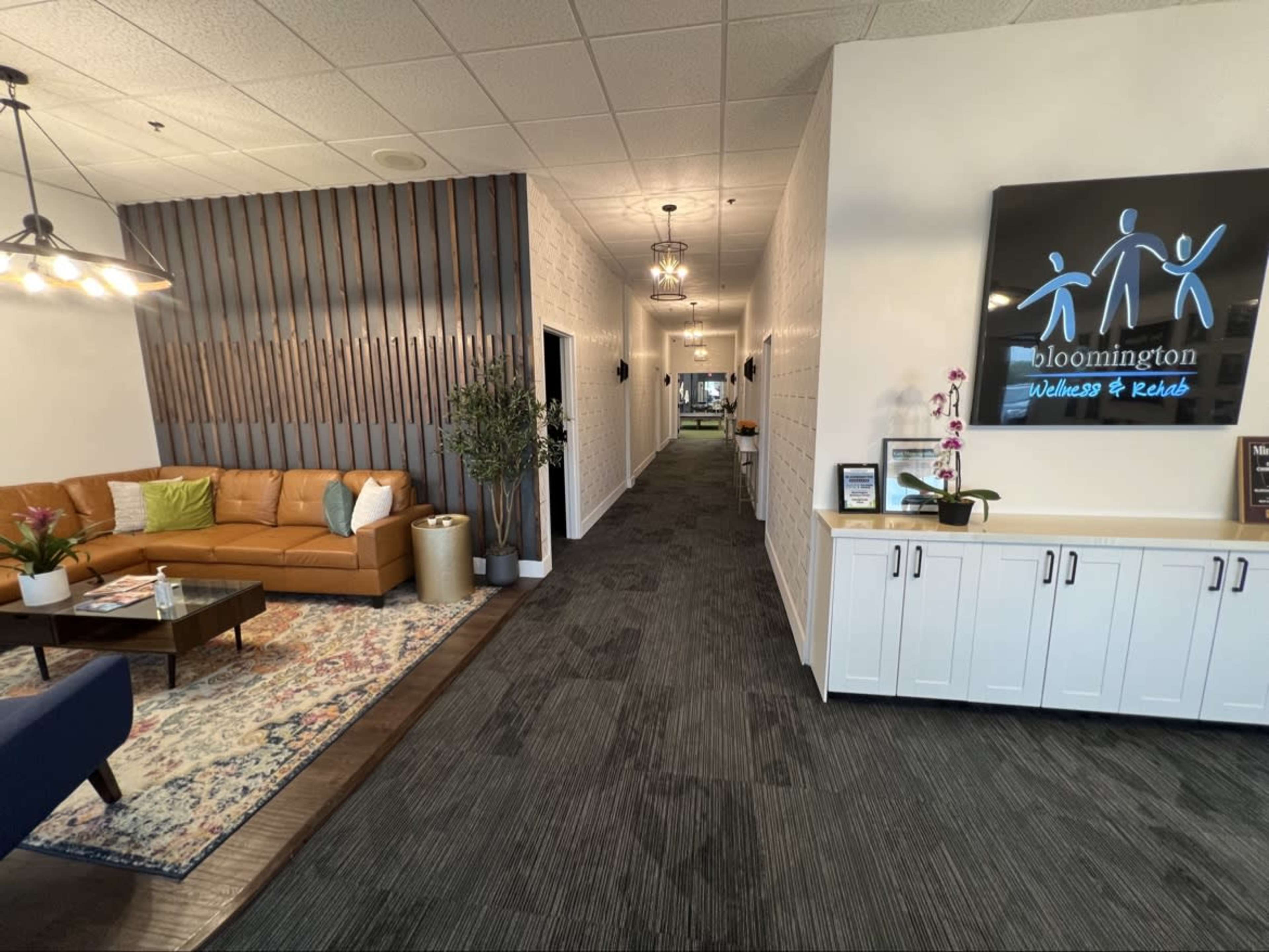 The image shows a modern, well-lit reception area with a brown leather couch, a coffee table, and a wall displaying the sign for "Bloomington Wellness & Kids," leading into a long hallway.