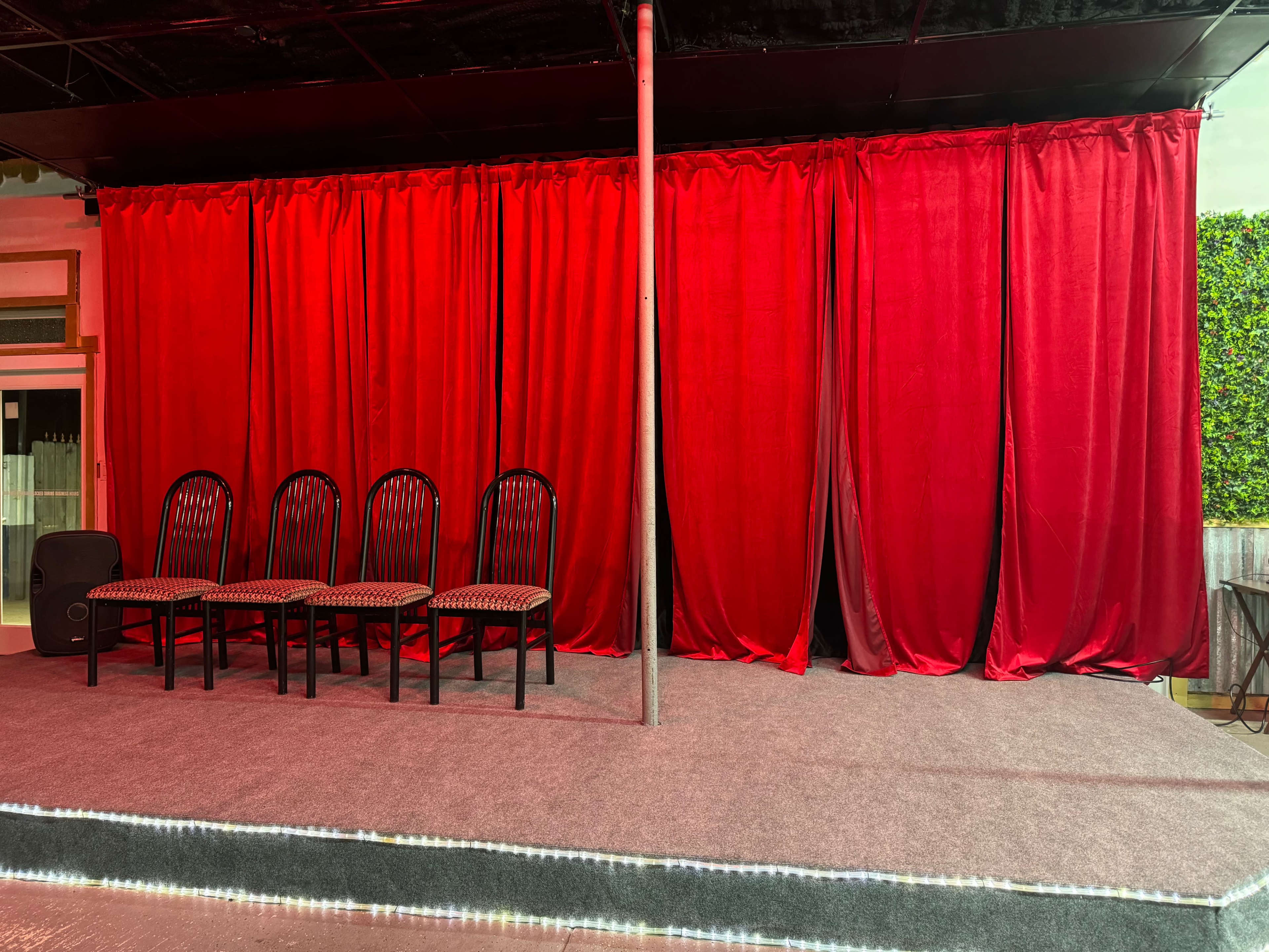 A stage with a gray carpet, six black chairs arranged in a row, and red curtains in the background.